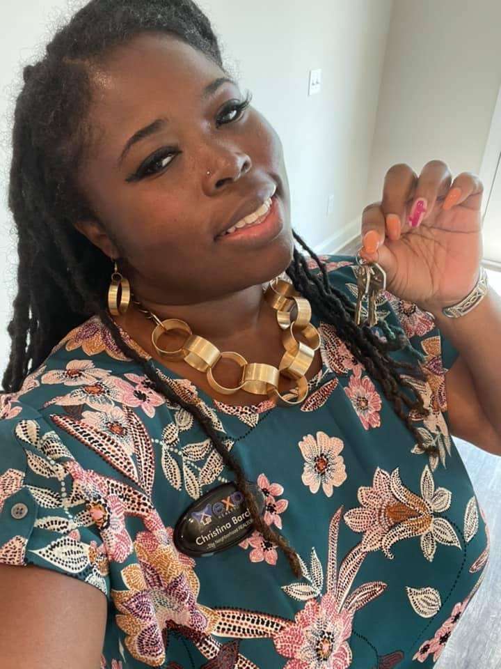 Woman with dark hair styled in dreadlocks, wearing a floral top, gold jewelry, and a black badge, smiling and holding a ring.