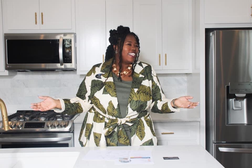 Woman with braided hair smiling and gesturing in a kitchen with white cabinets and stainless steel appliances.