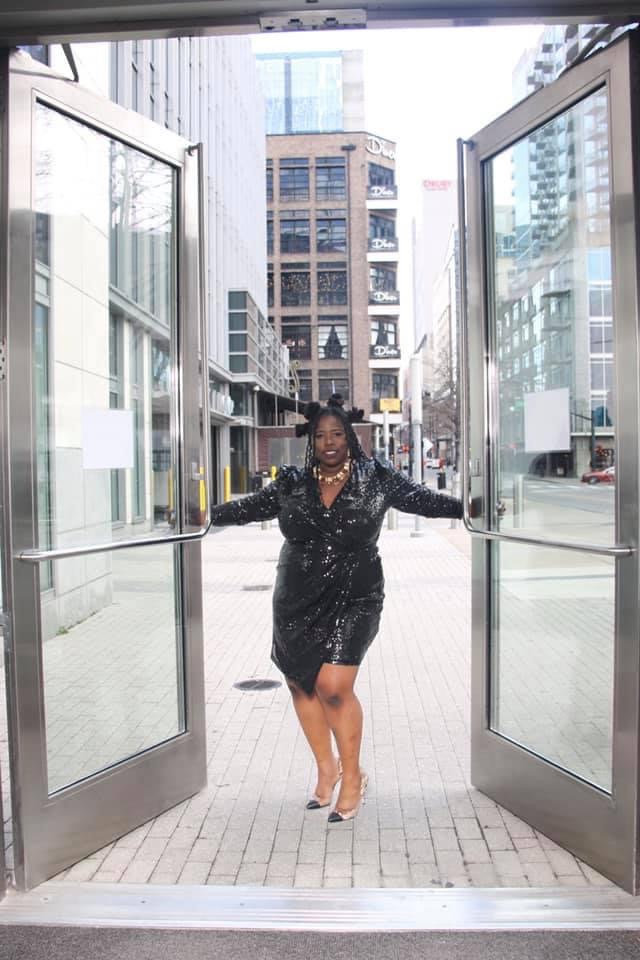 Woman in black dress standing in open glass doors with city buildings behind her.