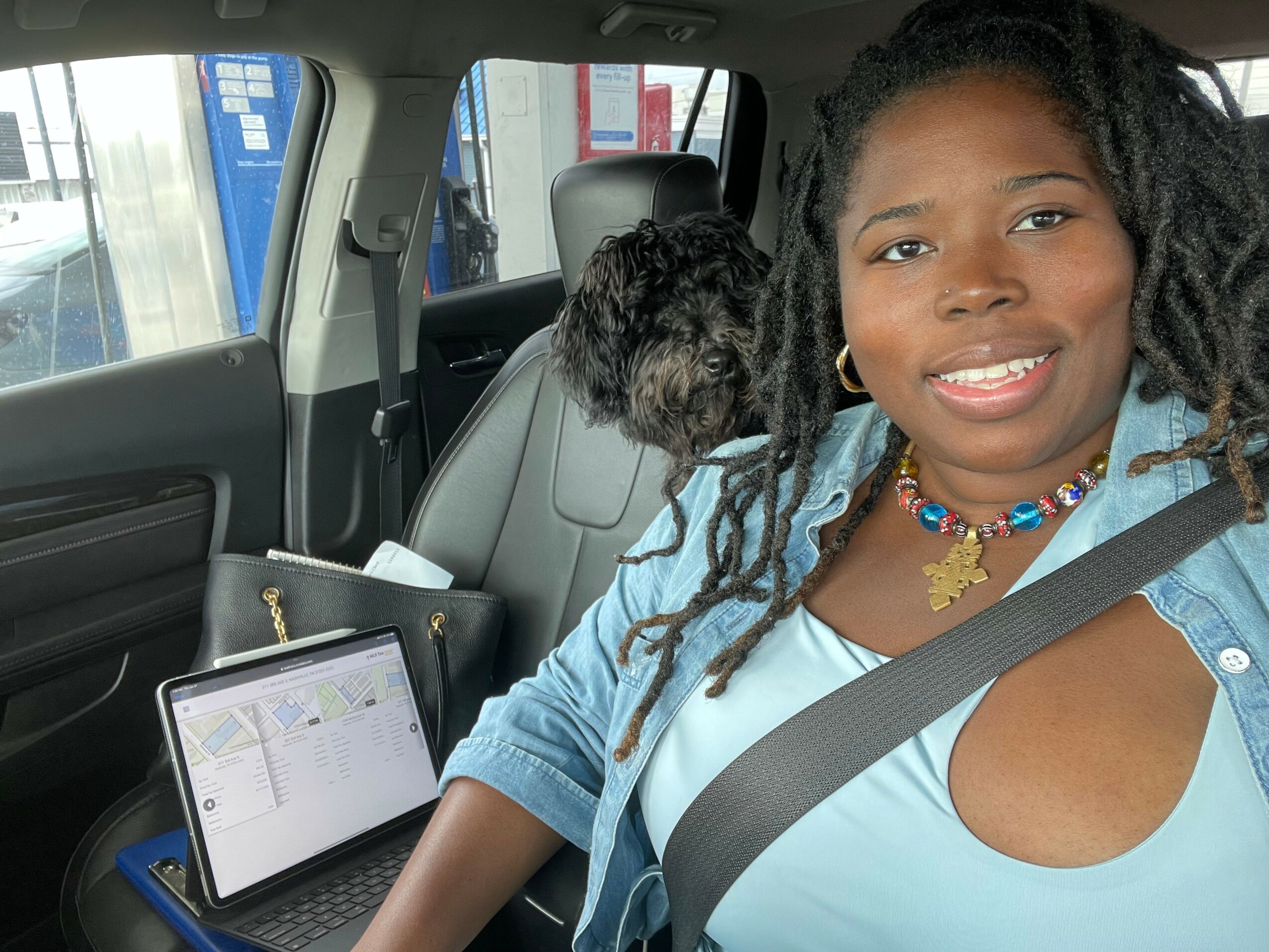 Young woman with braided hair smiling in car, wearing a blue top and colorful necklace, seatbelt on.