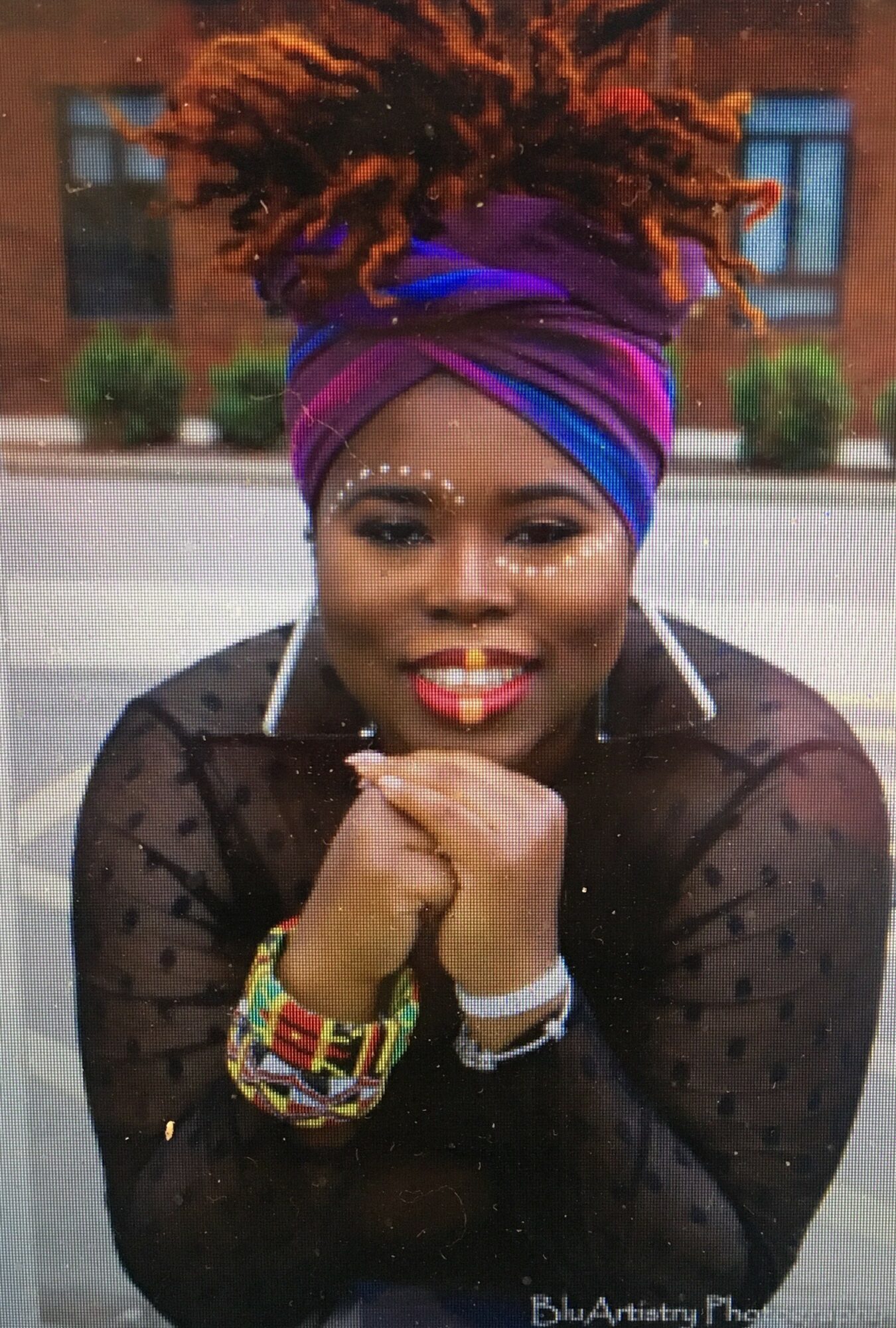 Woman with colorful headwrap, smiling, resting chin on hands, wearing earrings and bracelets, outdoors with buildings in background.