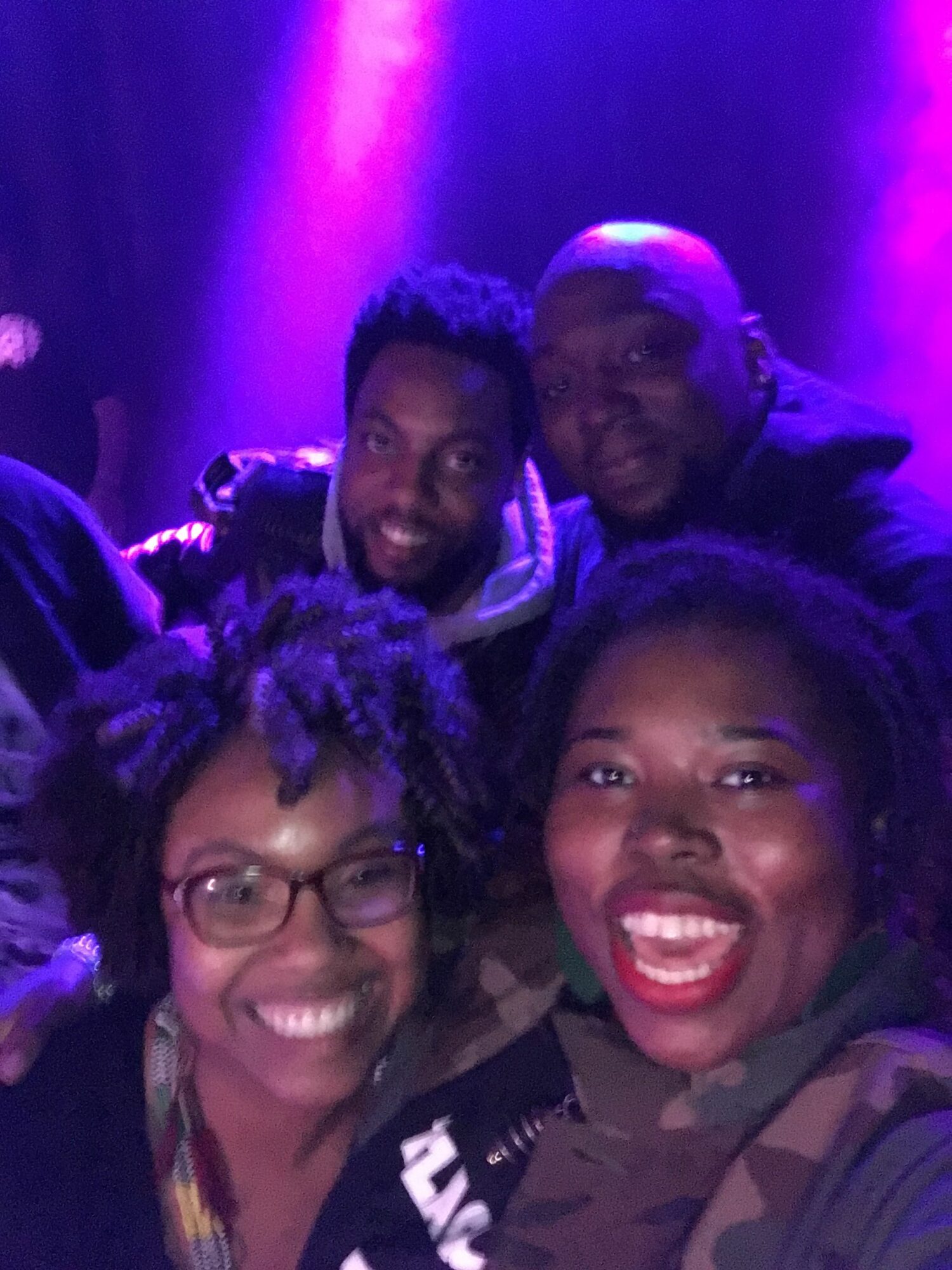 Four people smiling and posing together in a dark, colorful setting with purple lighting.