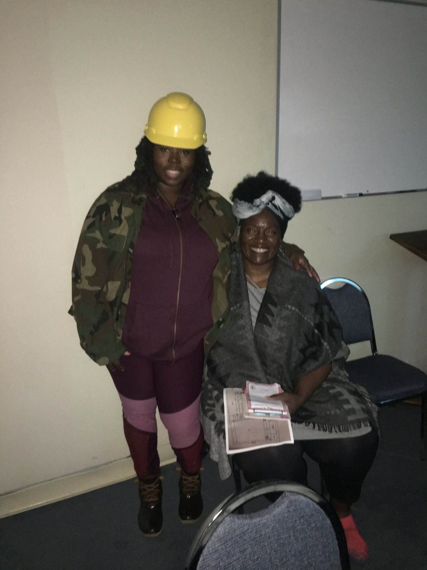 Two women standing indoors, one wearing a yellow helmet and camouflage jacket, the other holding a tablet, with chairs nearby.
