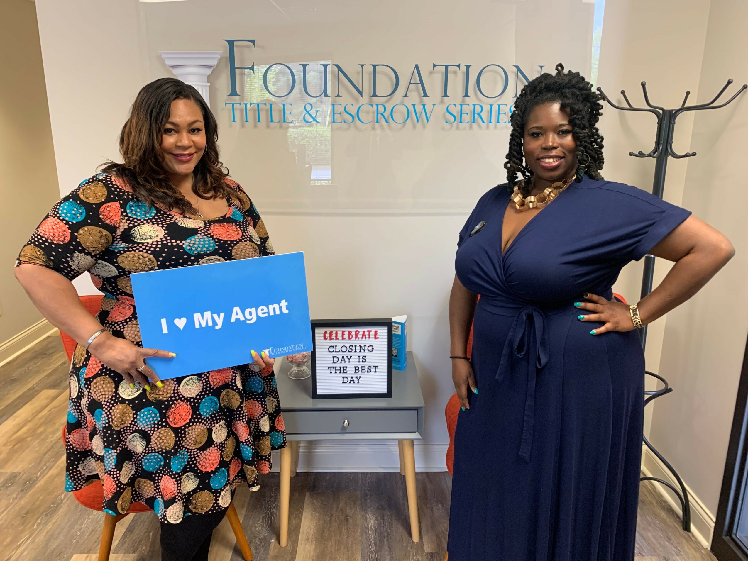 Two women stand in front of a sign that reads 'Foundation Title & Escrow Service.' One woman holds a blue sign that says 'I ❤️ My Agent.'