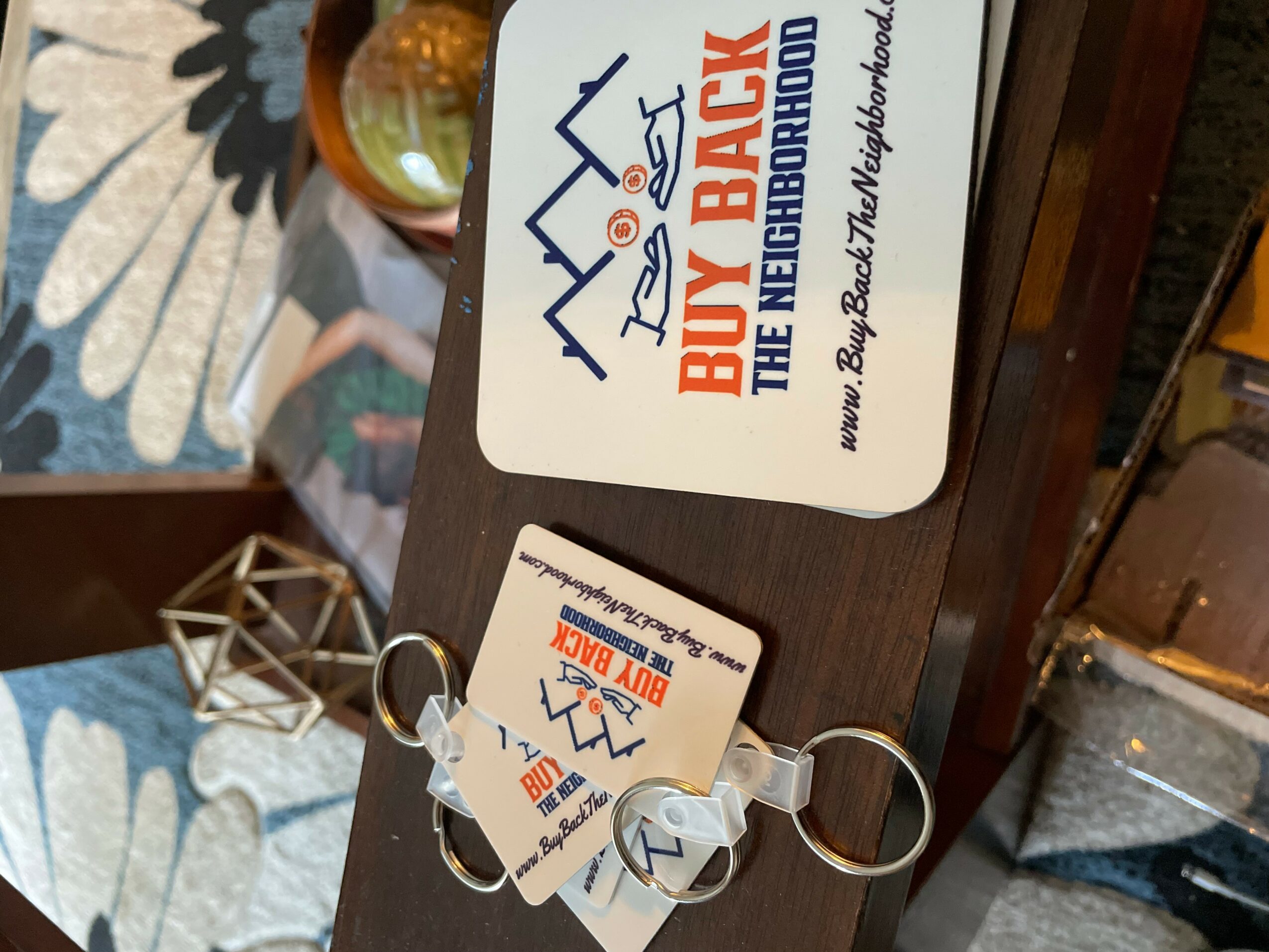 Business card with logo and text for Billy Raby, The Neighborhood, on a wooden surface, with keys attached, in a room with patterned carpet and furniture.