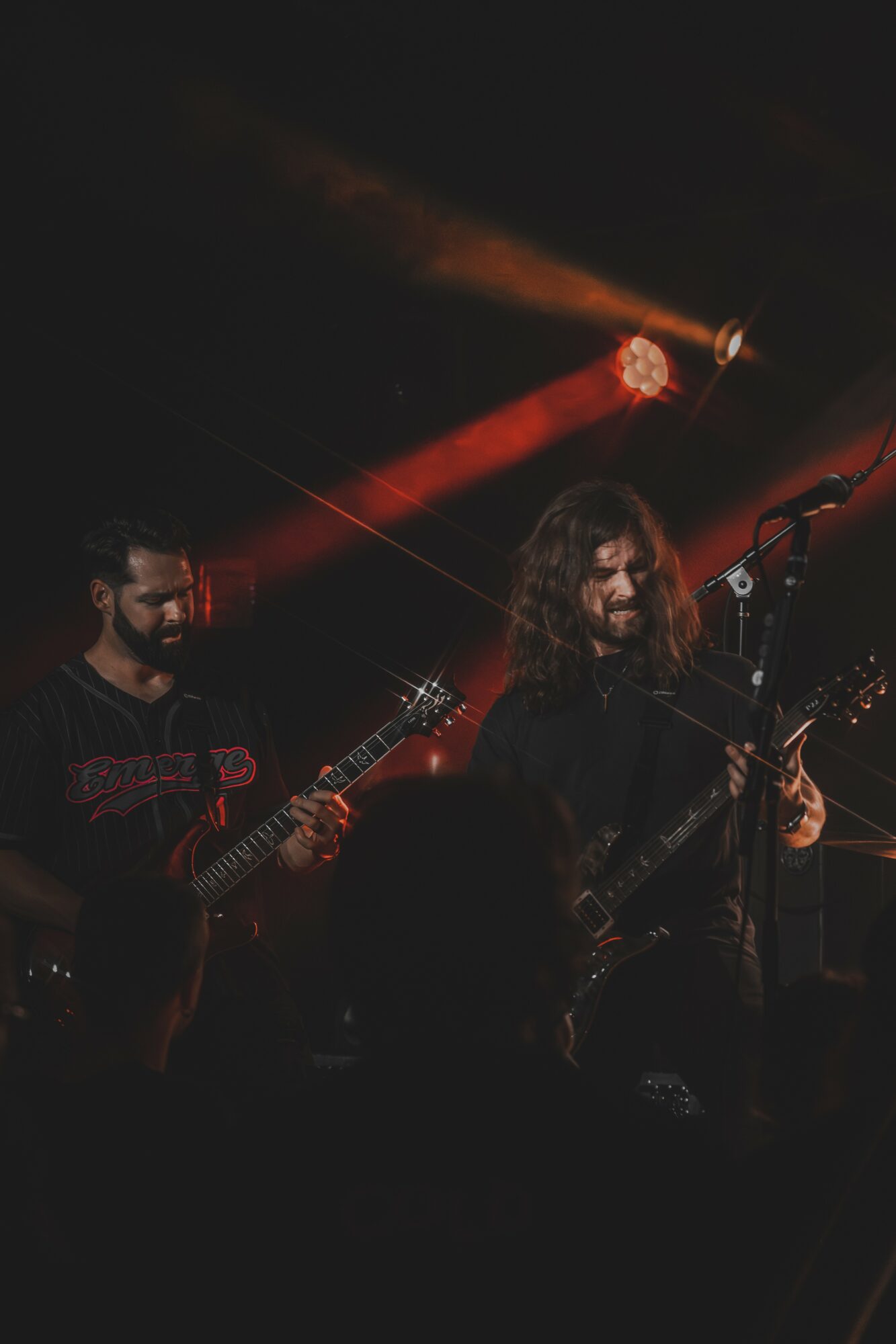 Two musicians playing guitars on stage with red and orange lighting, dark background, and audience silhouettes.