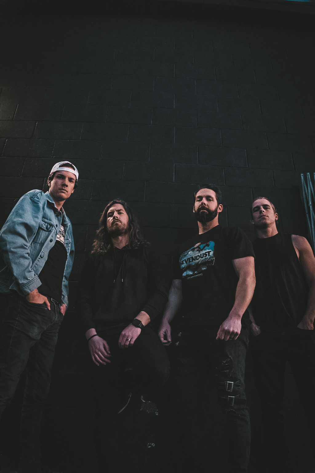 Four people standing against a dark wall, with one person wearing a denim jacket and cap, others in dark clothing.