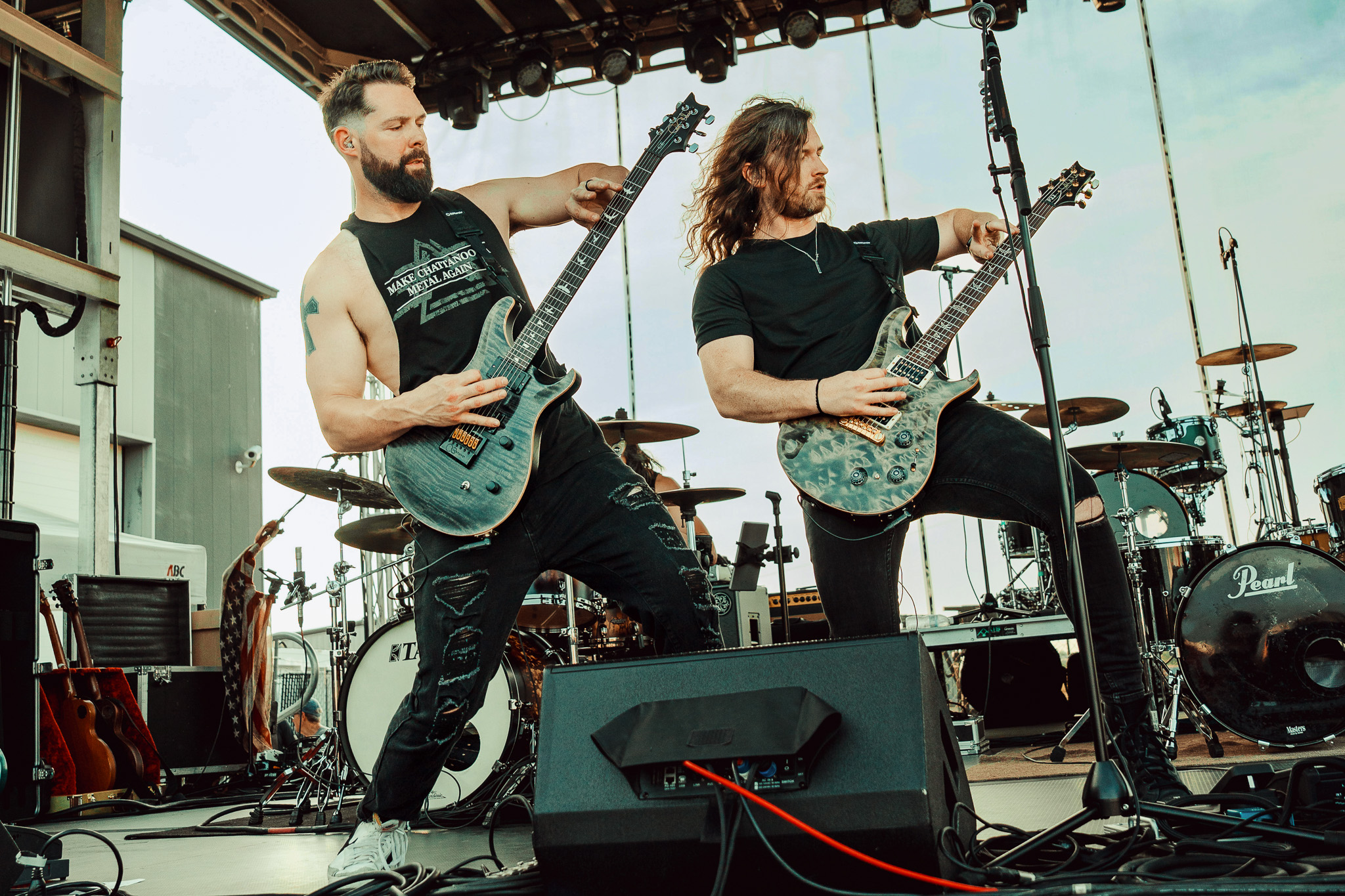 Two musicians playing electric guitars on stage with drums and equipment in the background, outdoors during daytime.
