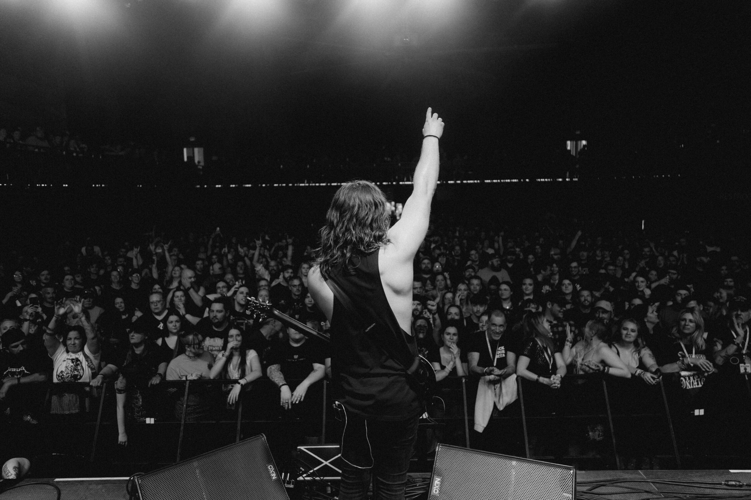 Person on stage with one arm raised, facing a large audience in a dark setting.
