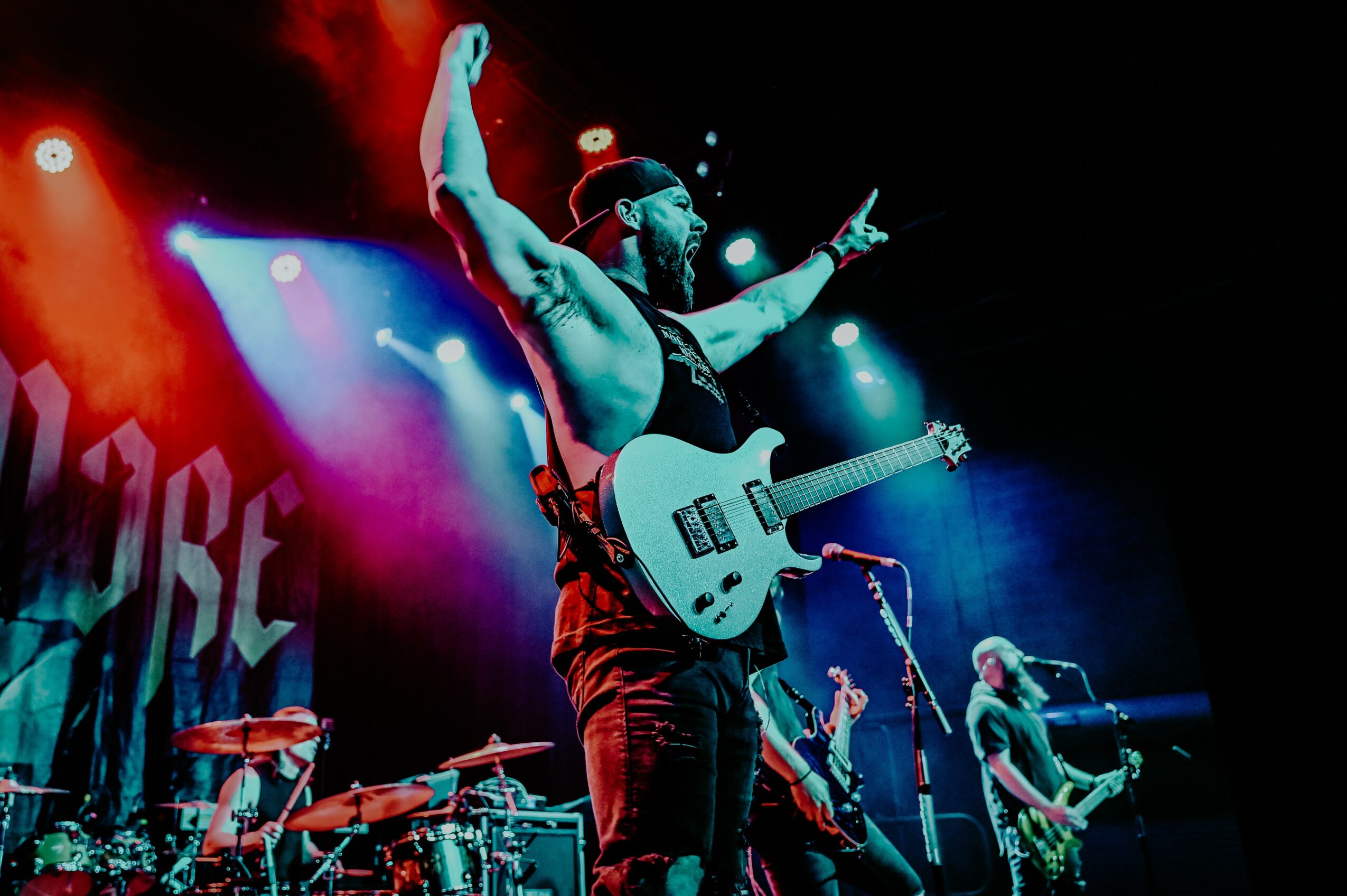 Musician on stage with guitar, raising arm, colorful lights, band members playing instruments in background.