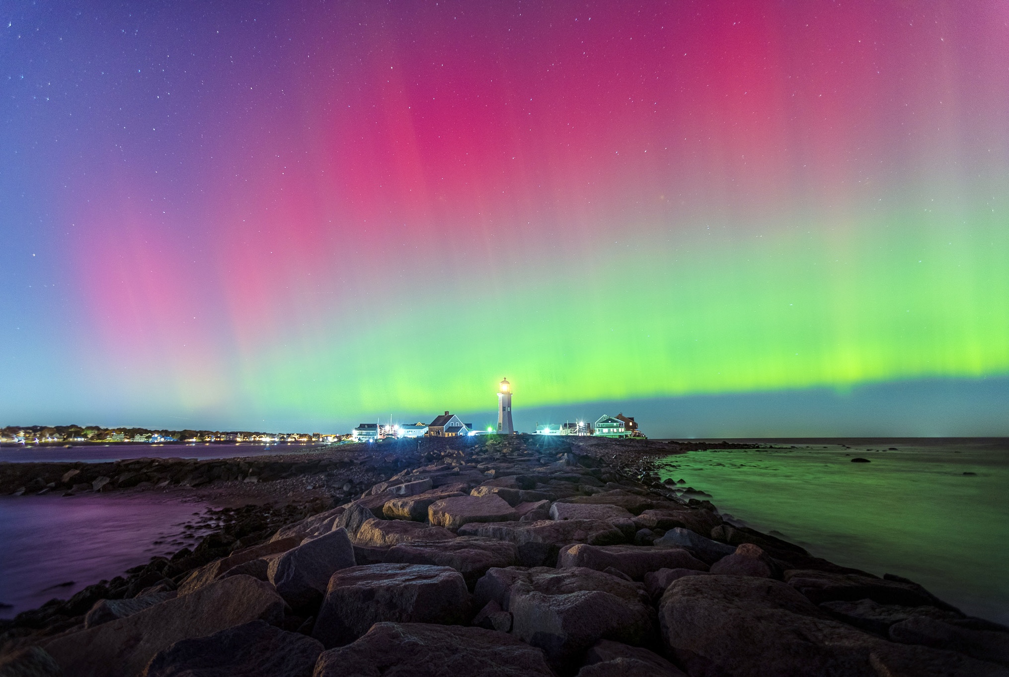Northern lights display colorful green, pink, and purple streaks over a rocky shoreline at night.
