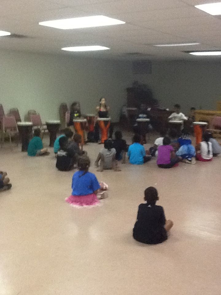 Children sitting on the floor watching a performance in a room with chairs and a piano, some children sitting alone, others in groups.