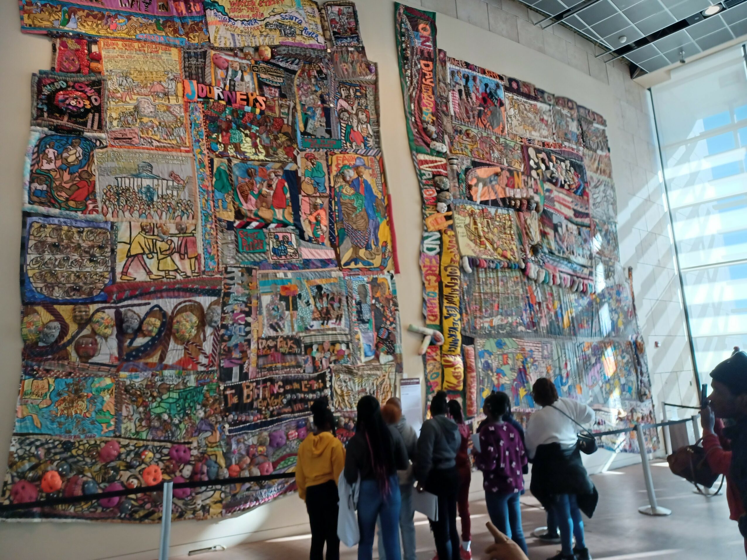 Group of people viewing large, colorful textile artworks displayed on a wall in a modern gallery space.