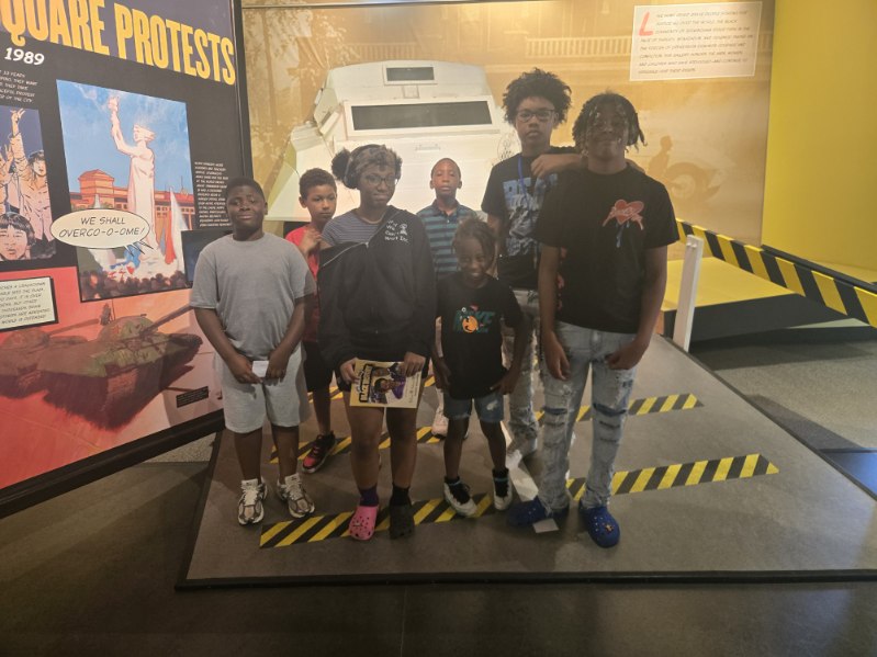 Group of seven children and teenagers standing in front of a display at a museum, with a yellow wall and caution tape nearby.