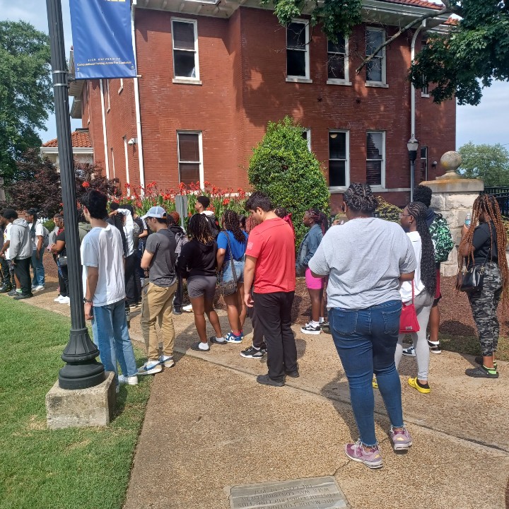 Group of people standing outside a brick building with trees and a sidewalk, some waiting in line or talking.