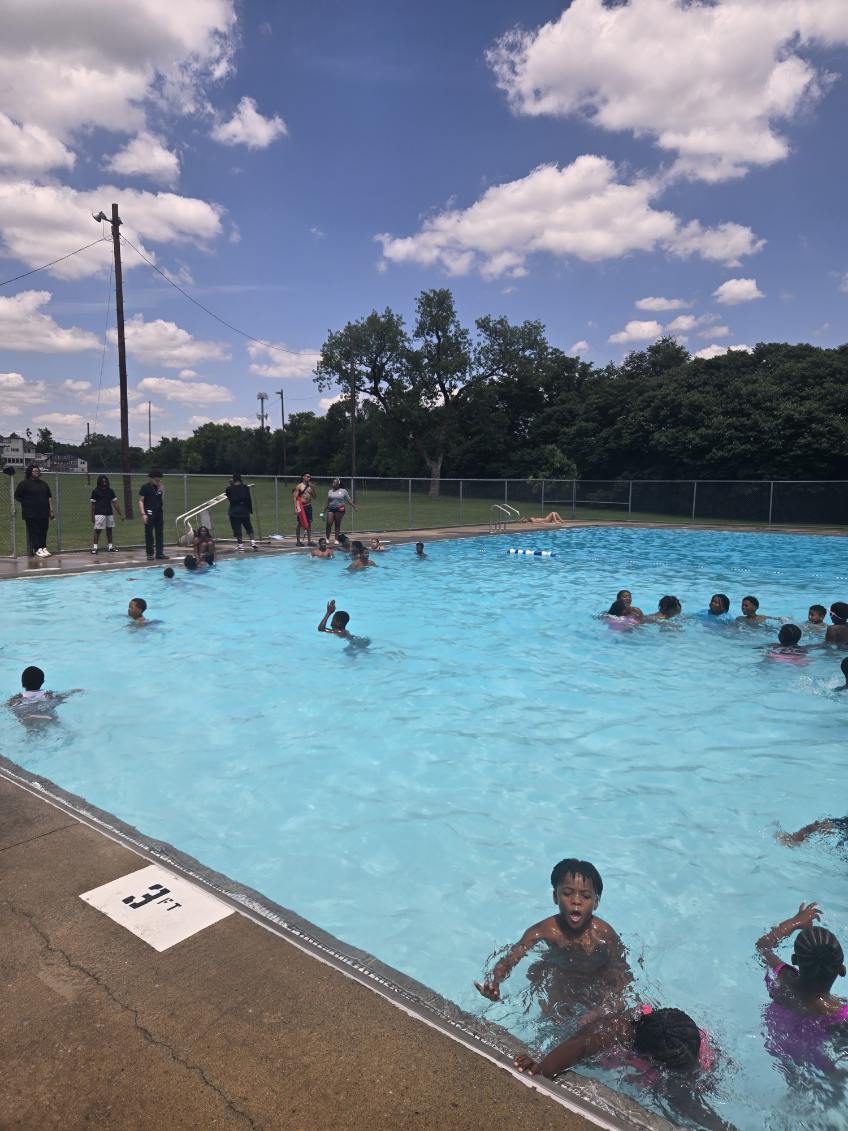 Children swimming and playing in an outdoor pool on a sunny day with clouds and trees in the background.