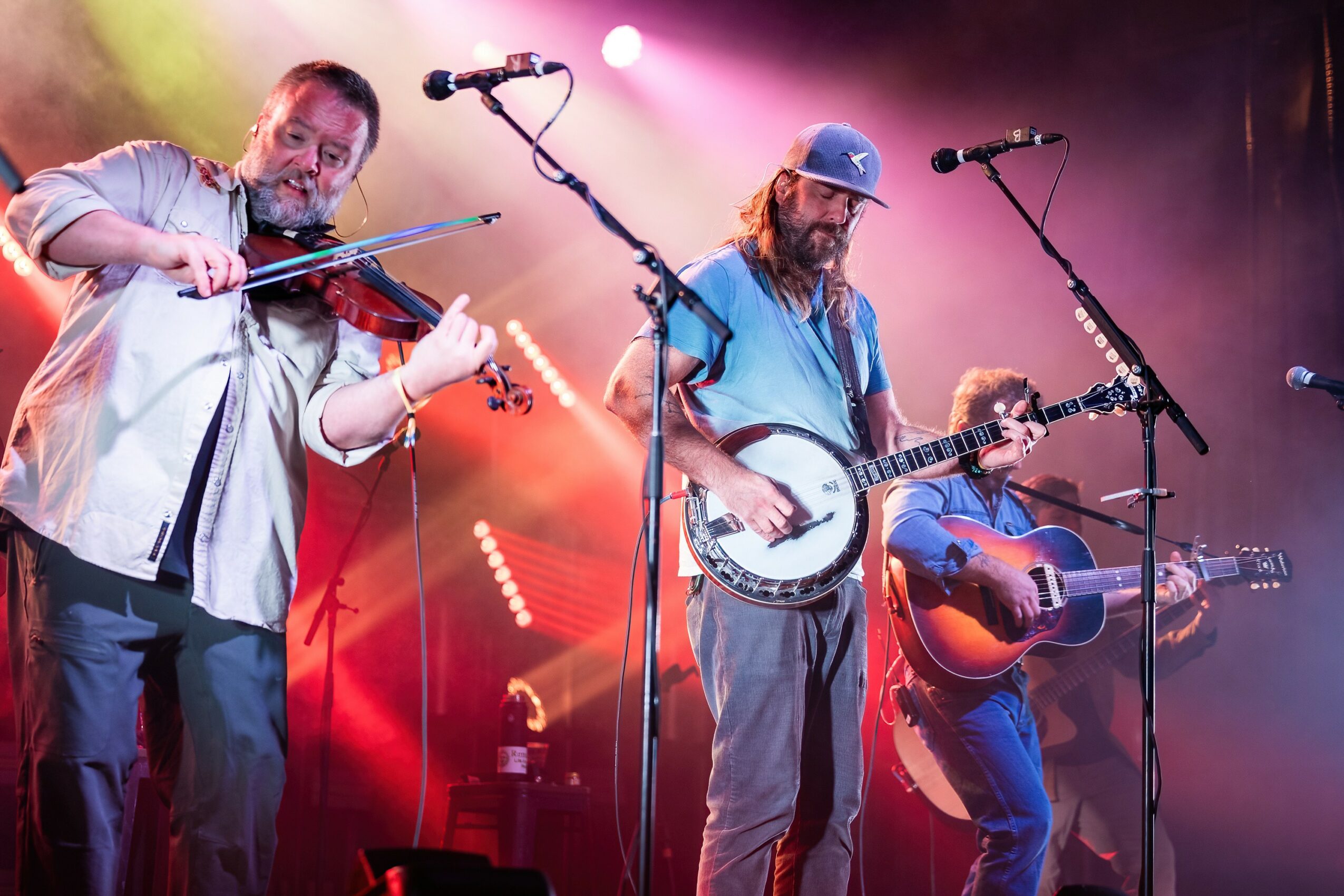 Three musicians perform on stage with colorful lighting, one playing violin, another with a banjo, and the third with a guitar.