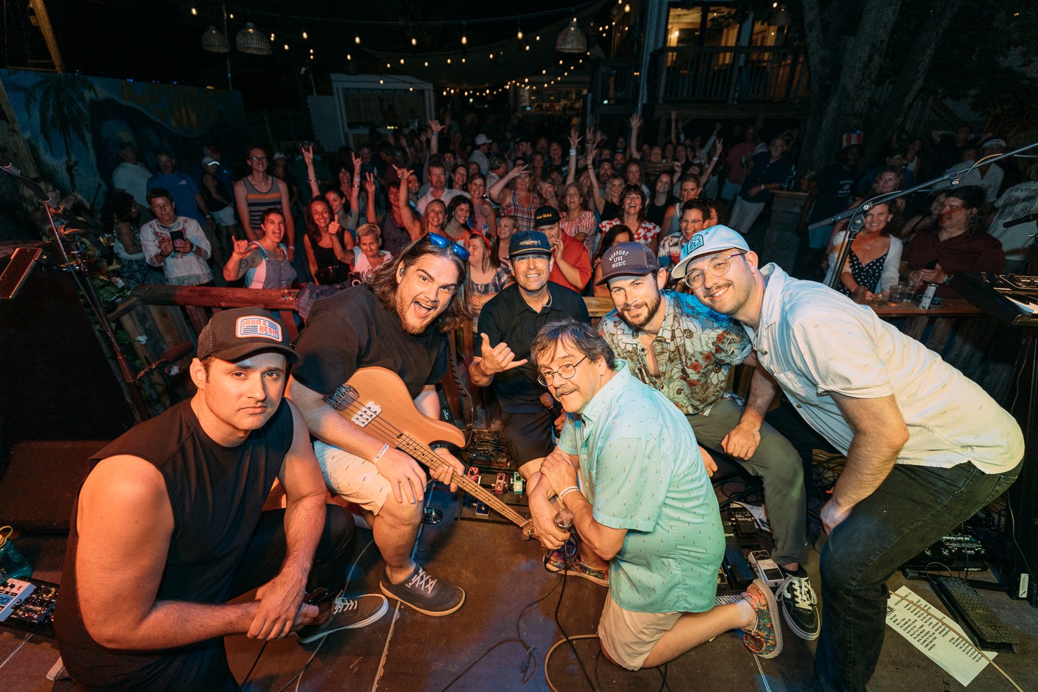 Group of six people posing with a guitar at an outdoor event during nighttime, with a crowd in the background.