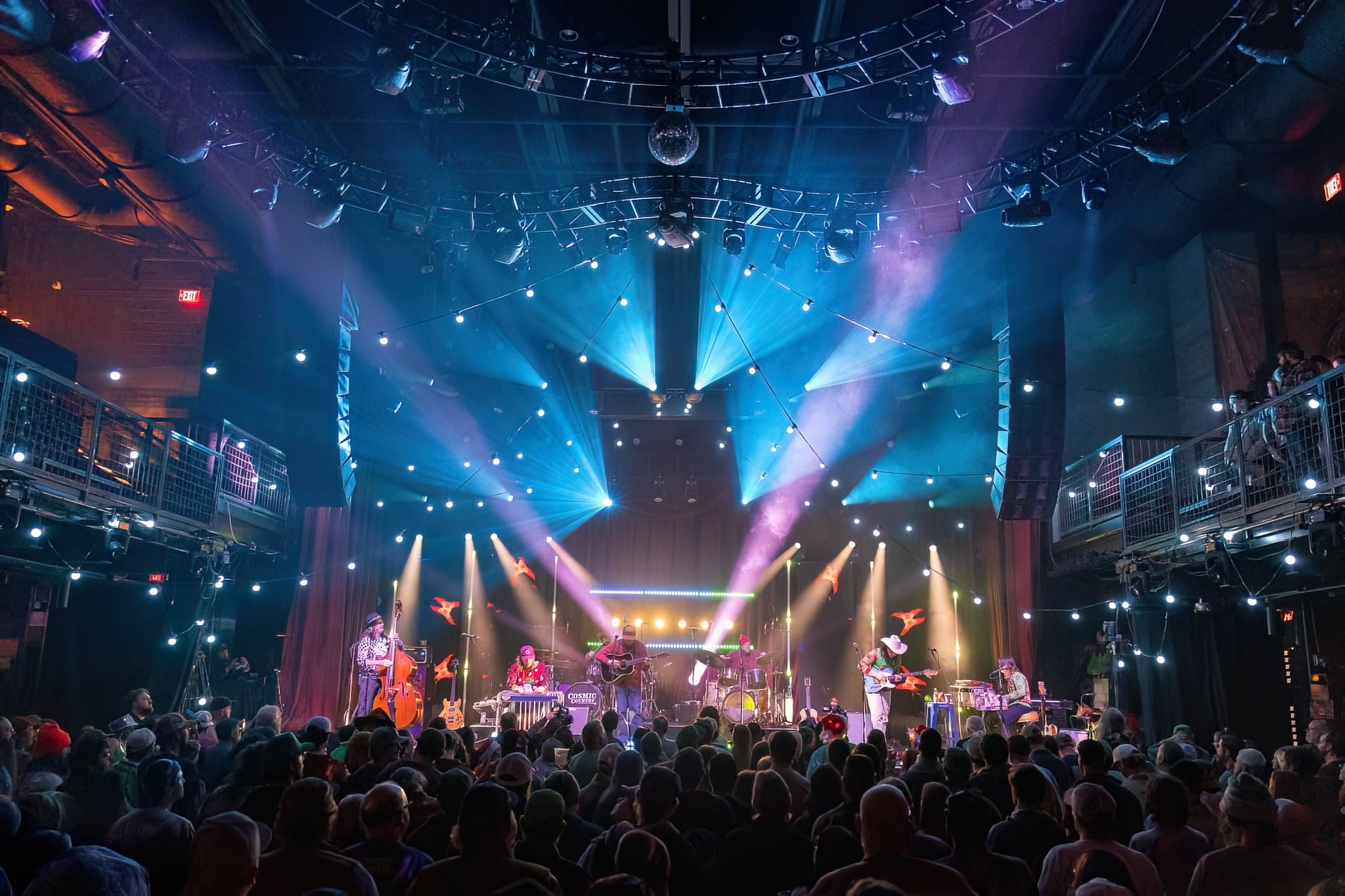 Concert stage with colorful lights and audience, performers on stage, in a large indoor venue with balconies.