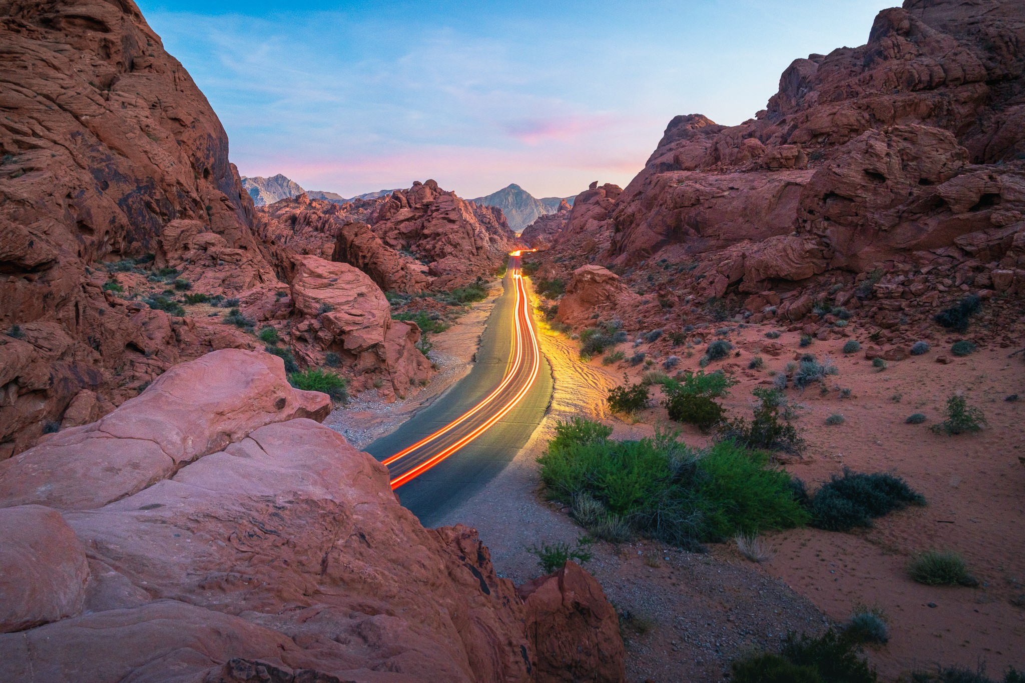 A winding road through a desert landscape with reddish rocky hills and sparse green vegetation, under a partly cloudy sky.
