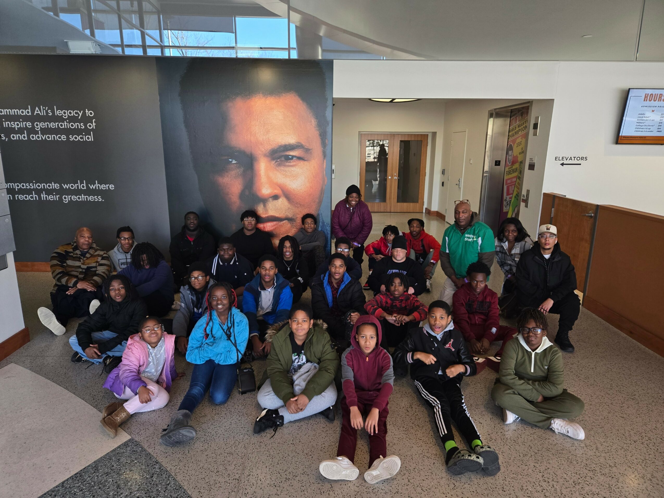 Group of children and adults posing in front of a large portrait of a man in an indoor setting.