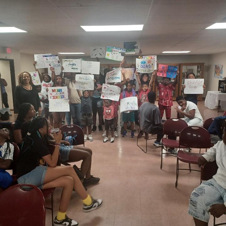 Group of children and adults holding signs in a room with chairs and artwork on walls.