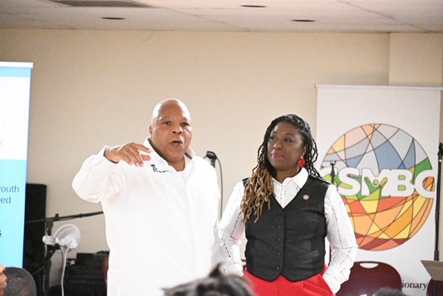 Two people standing in front of a whiteboard, one speaking and gesturing, the other listening, in a room with a presentation screen.