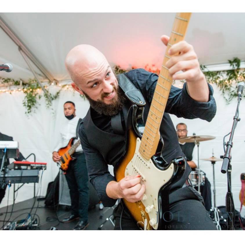 Bald man with a beard playing an electric guitar on stage, with band members and musical equipment in background.