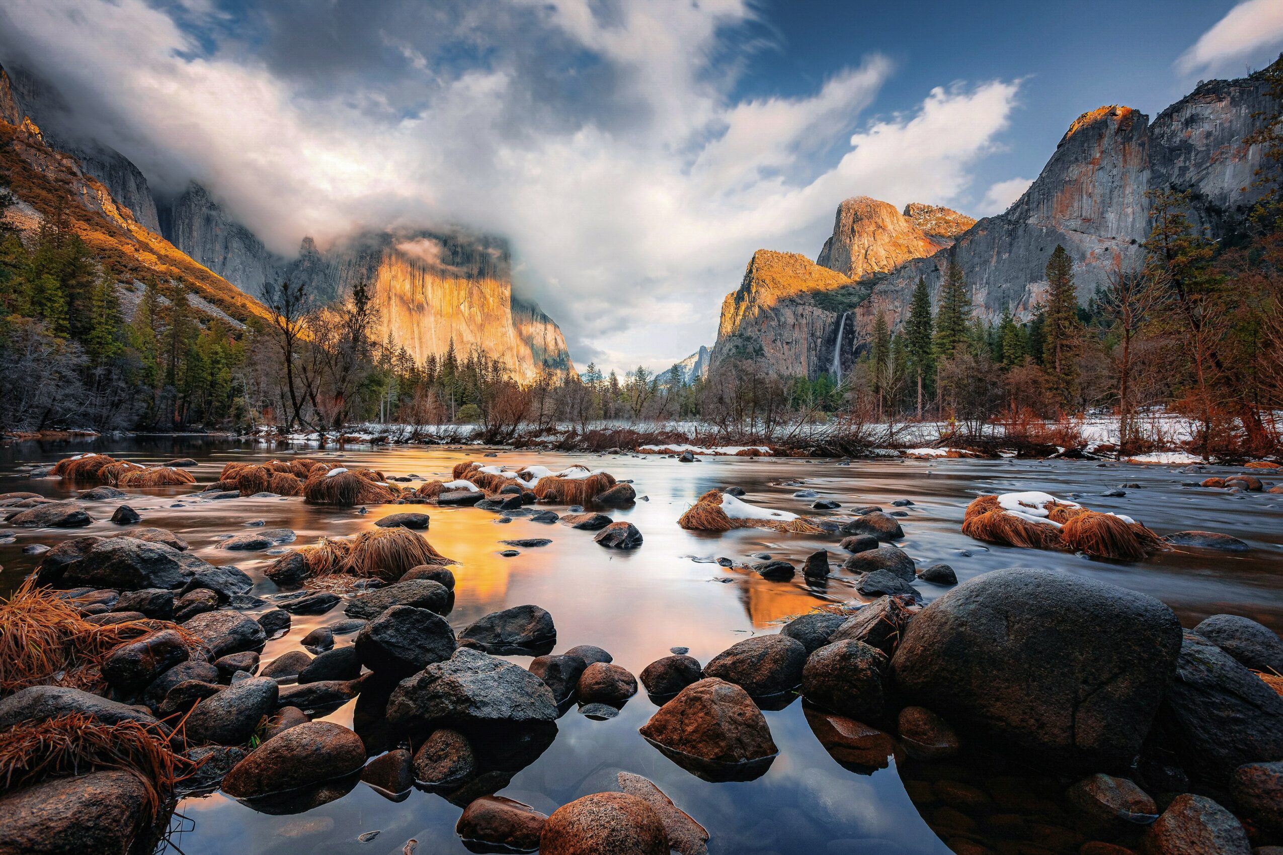 Scenic landscape of a river with rocks, surrounded by mountains and trees under a partly cloudy sky.