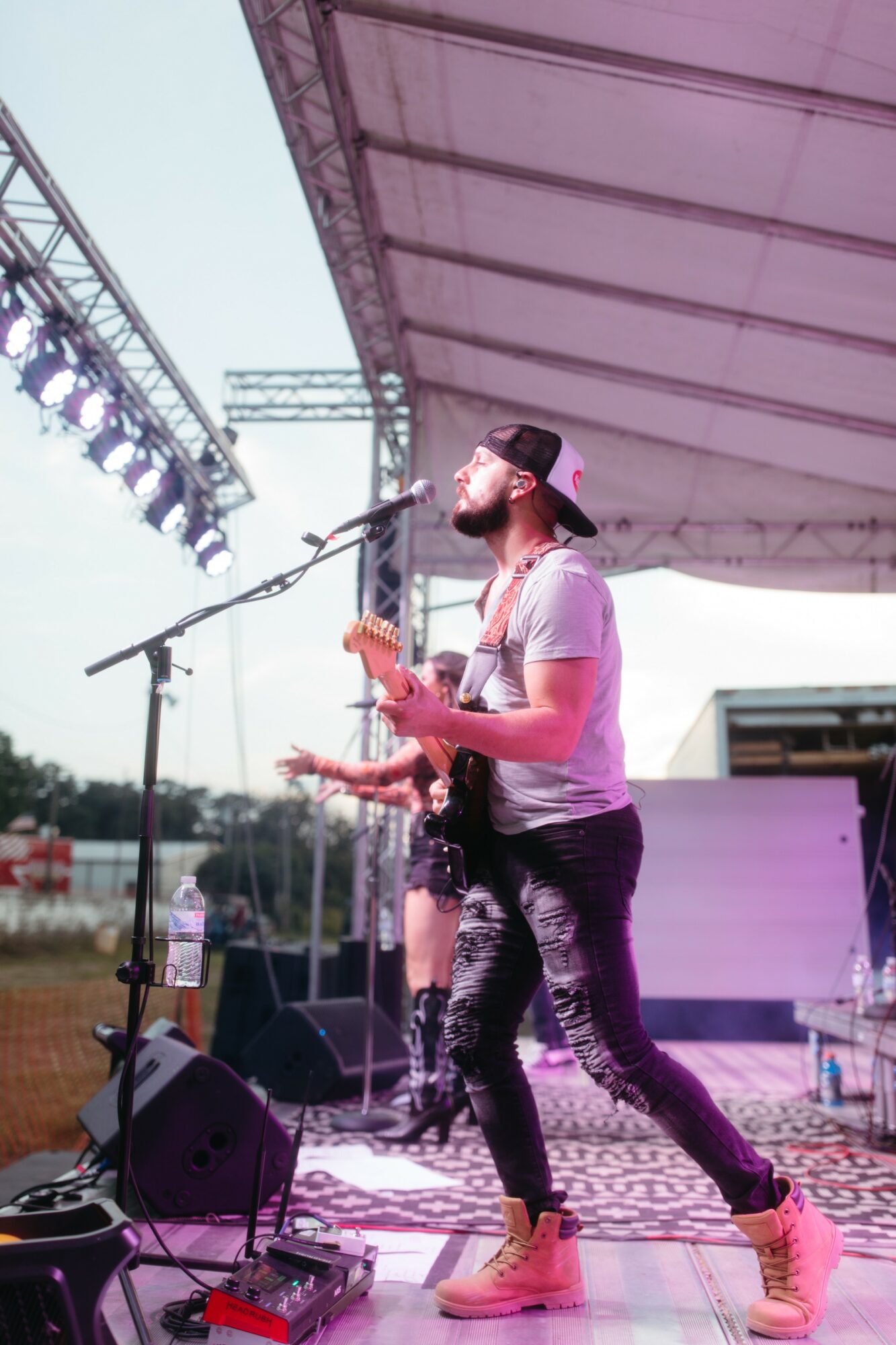 A person with a beard and cap performs on stage with a guitar, microphone, and stage equipment visible.