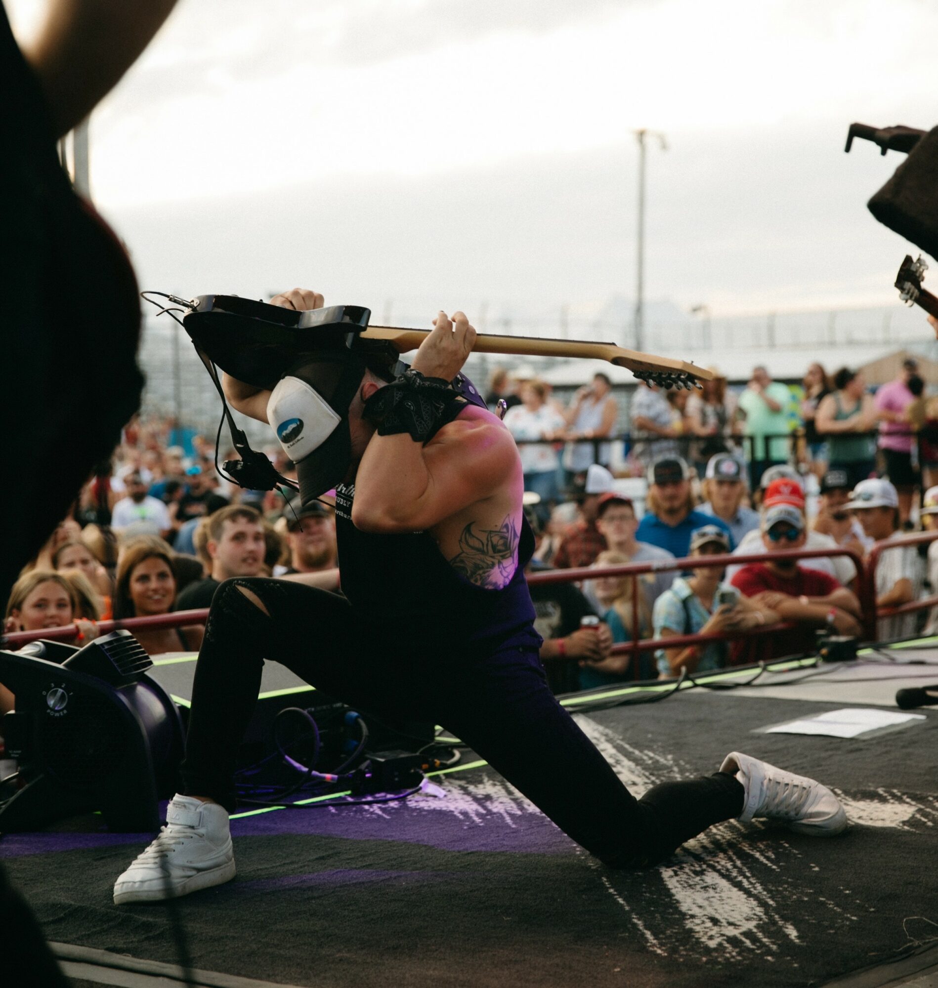 Musician performing on stage with a crowd in the background, holding a guitar over their shoulder, wearing headphones and white sneakers.