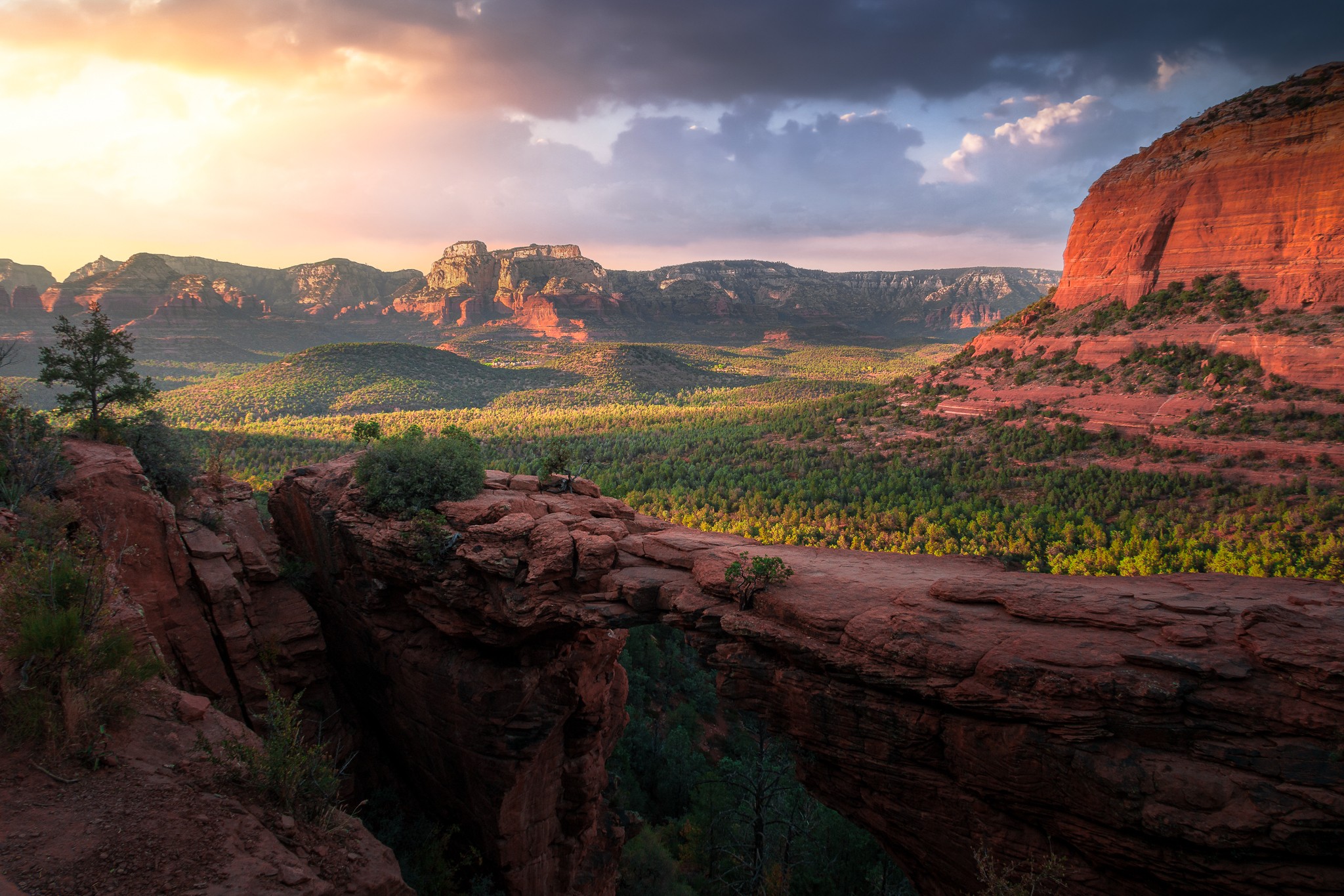 Landscape with red rock formations, green vegetation, and a partly cloudy sky at sunset.