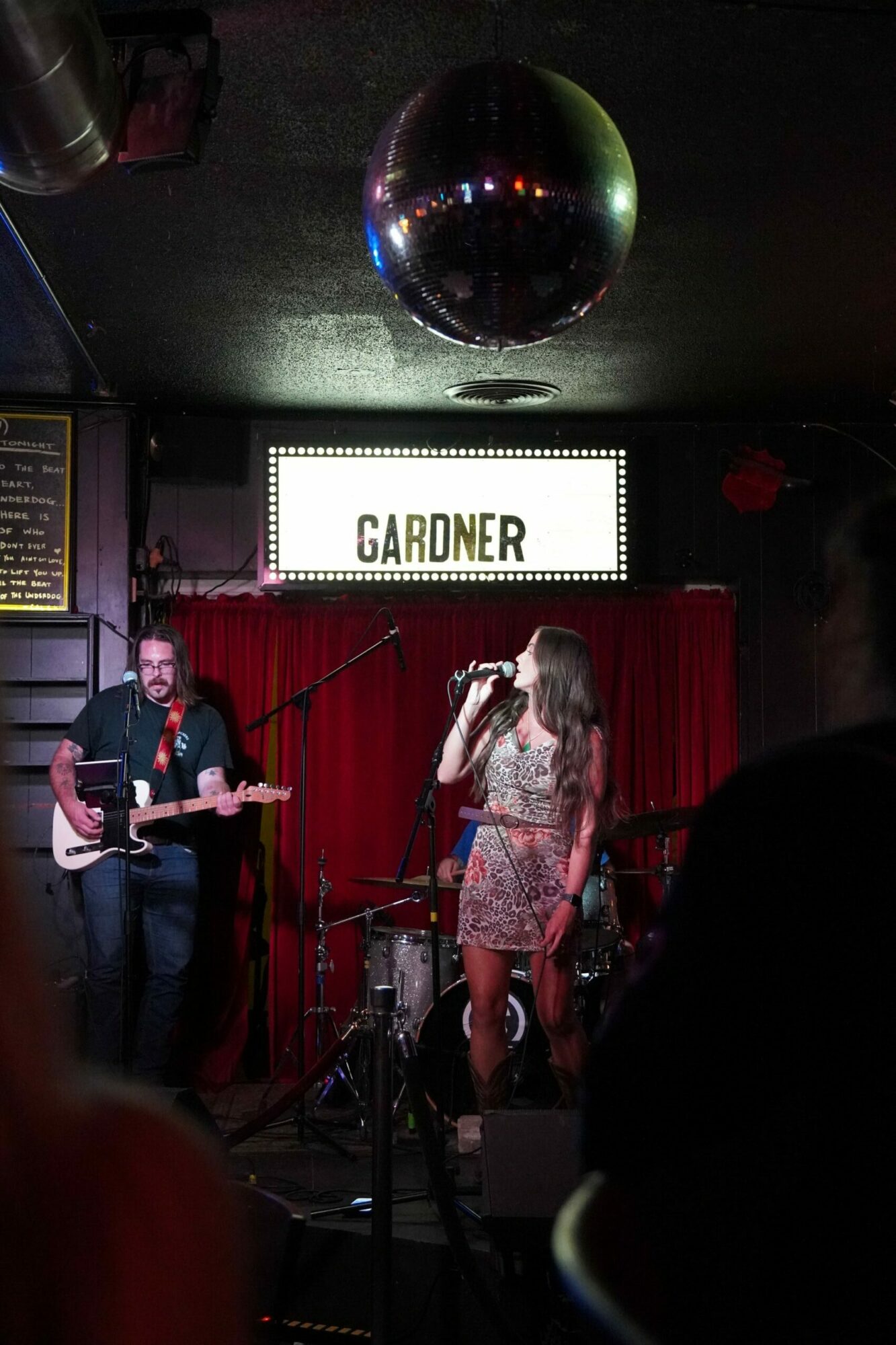 Two performers on stage with a red curtain background, one playing guitar and the other singing, with a sign reading 'GARDNER' behind them.