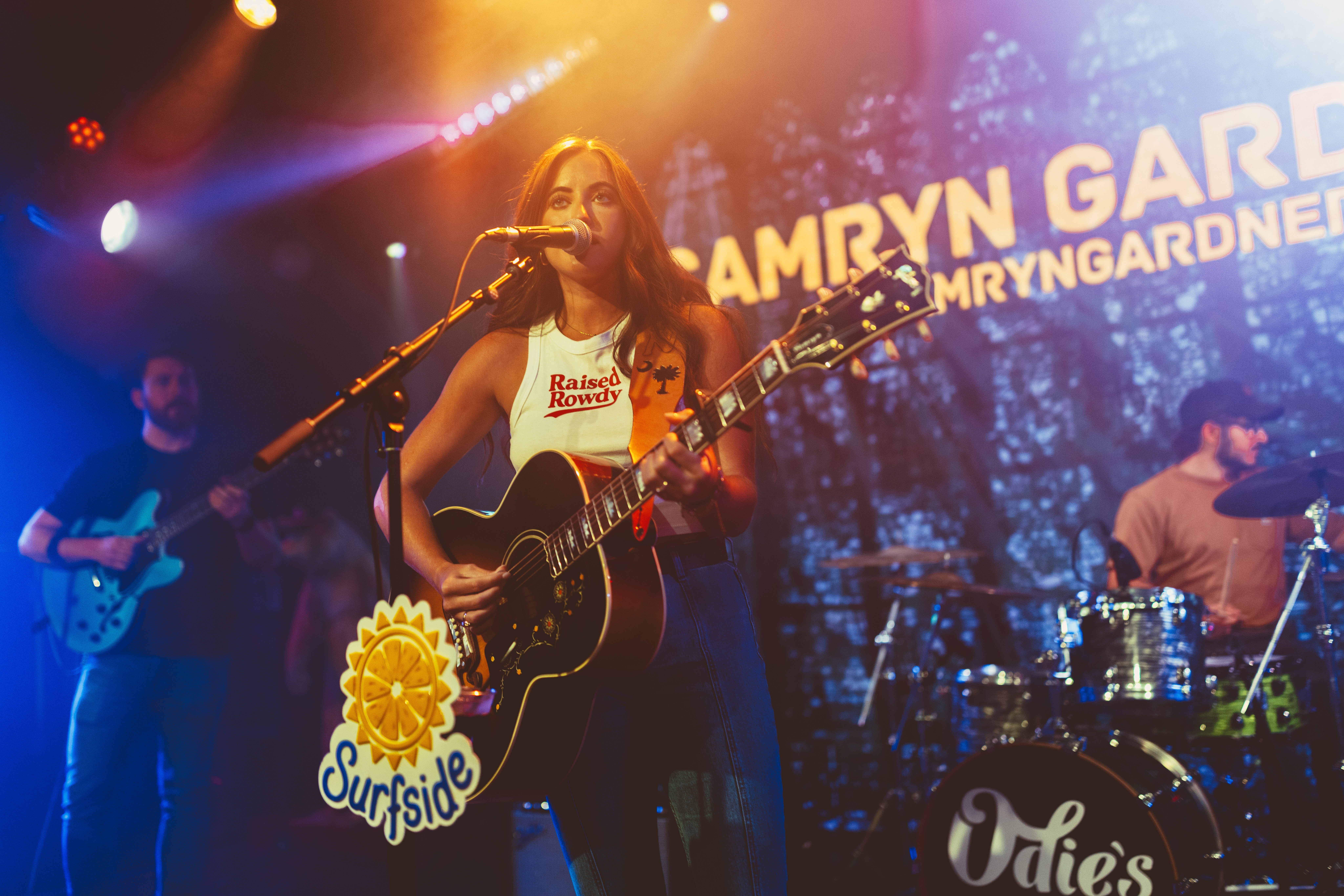 Female musician playing guitar on stage with band, colorful lights, and a large screen in background displaying text.