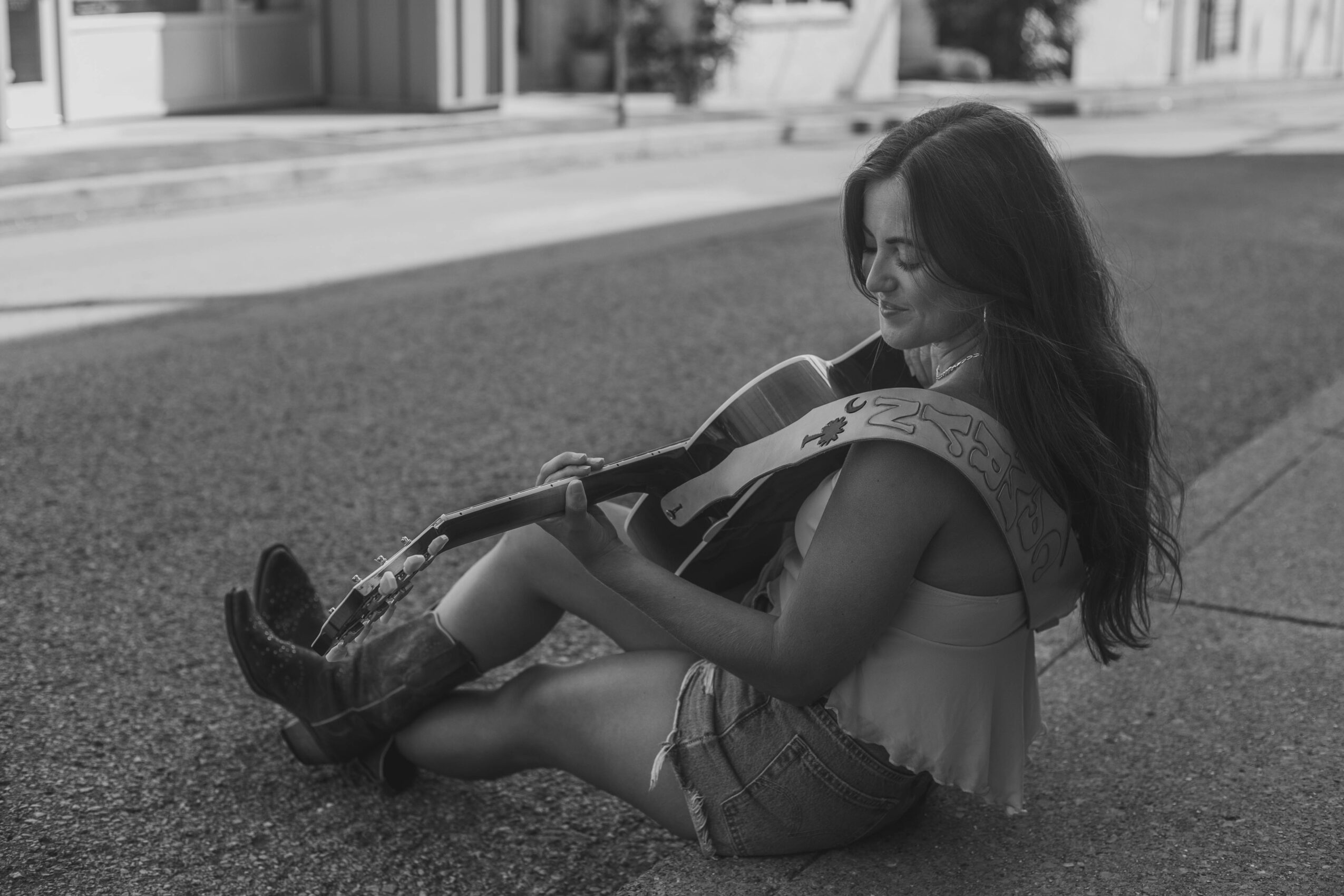 Girl sitting on the sidewalk playing an acoustic guitar, smiling, with long hair and casual clothing, in black and white.