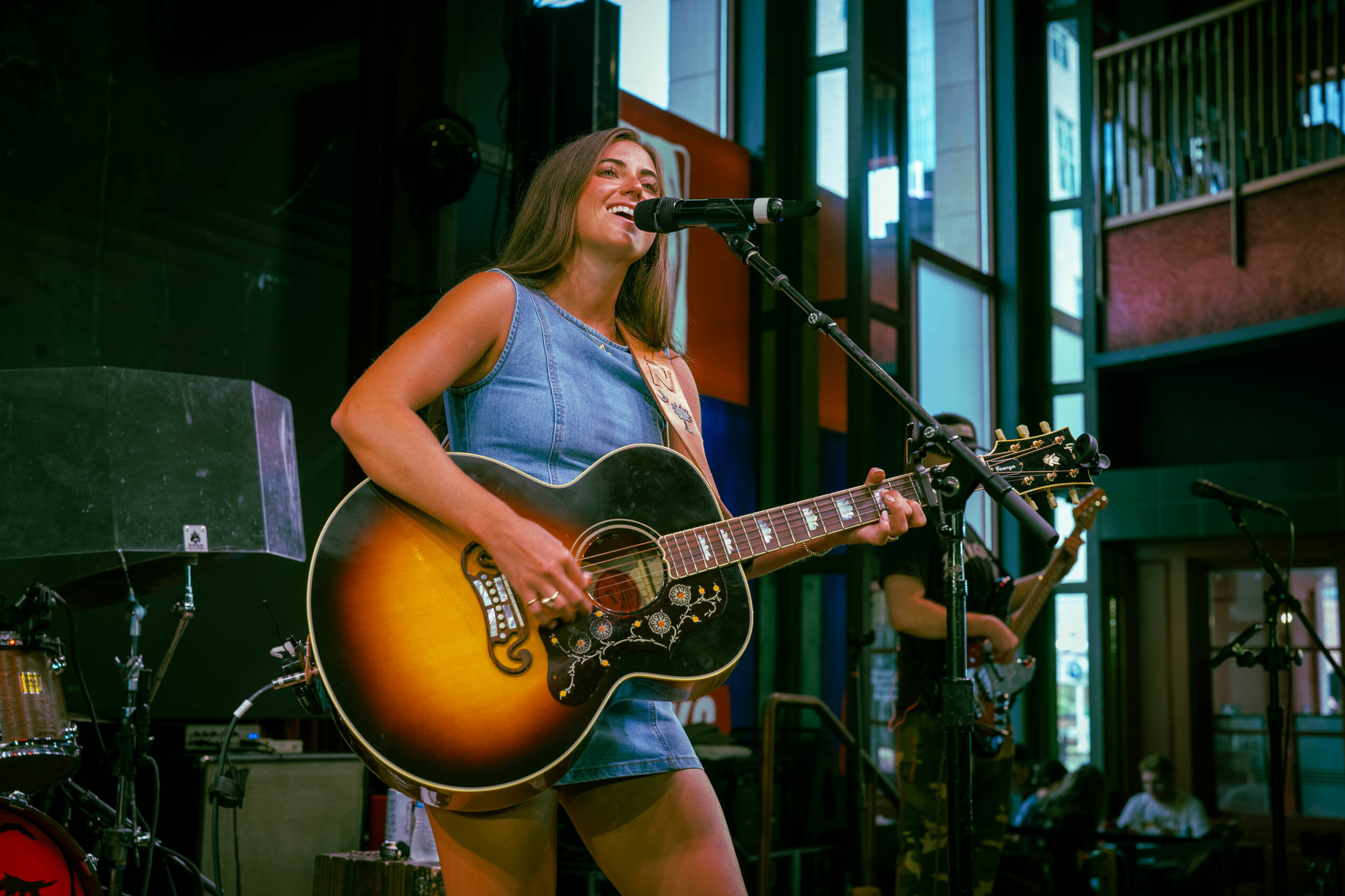 Woman singing and playing guitar on stage with microphone, smiling, in a modern indoor venue.
