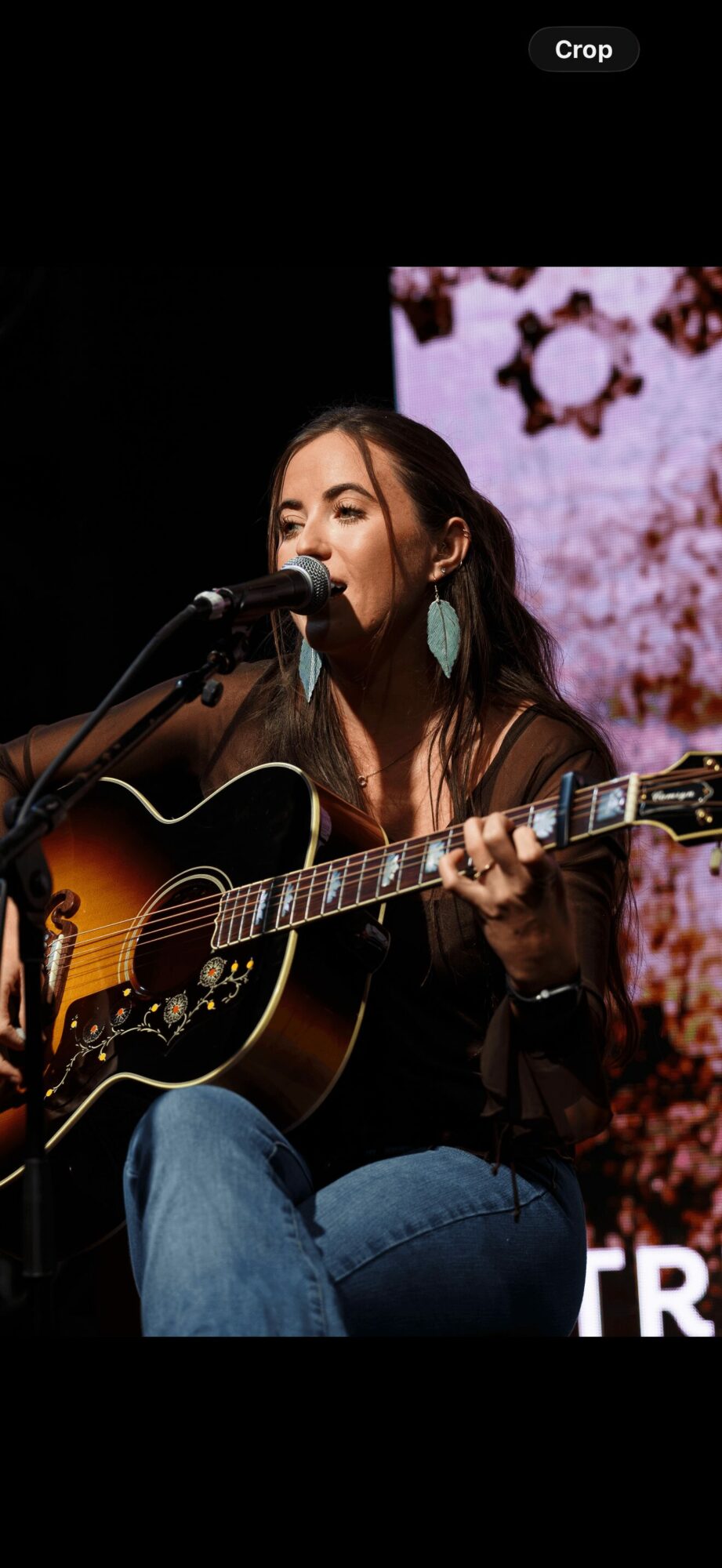 Woman playing guitar and singing into a microphone on stage with a pink floral background.