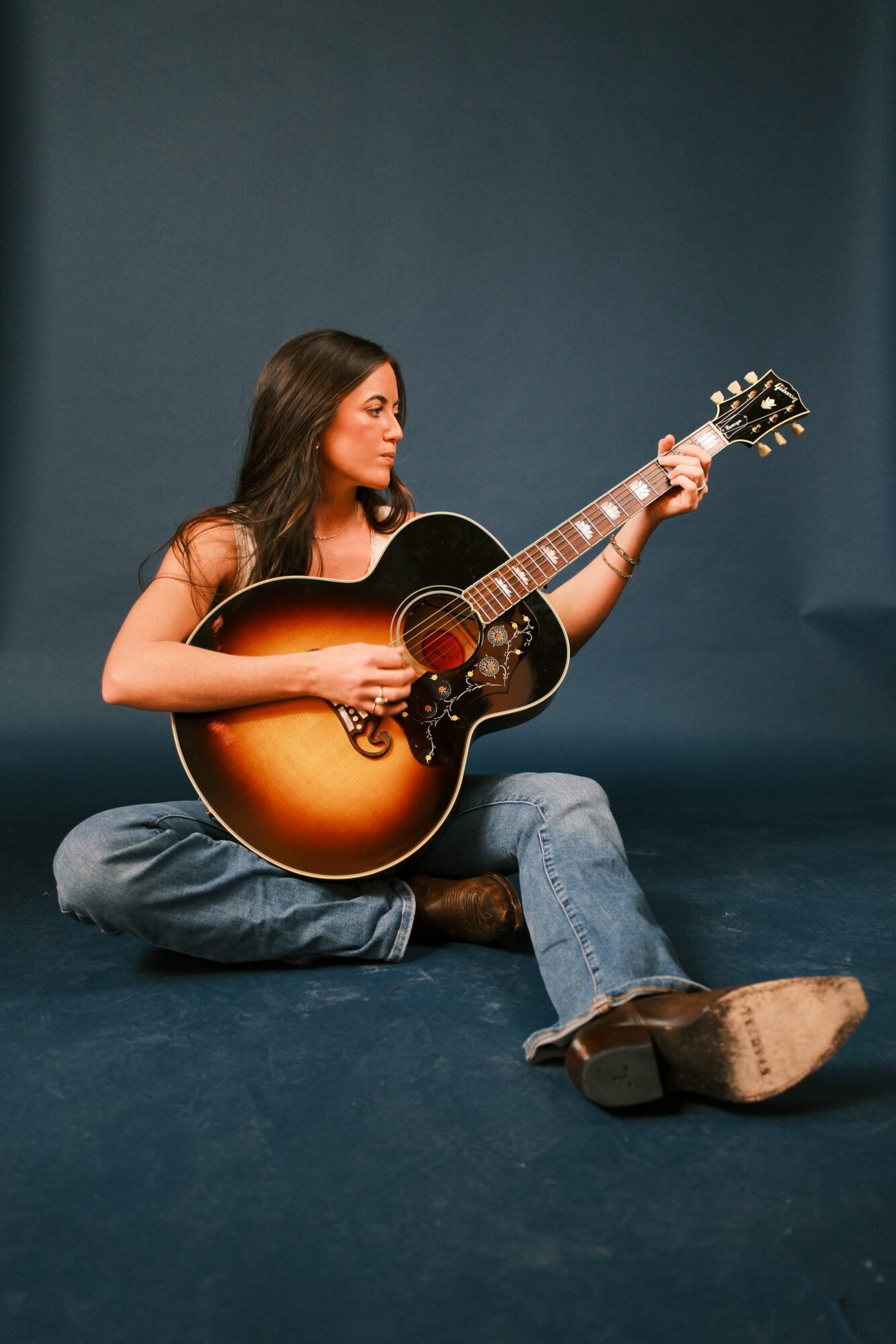 Woman sitting on the floor playing an acoustic guitar, with a plain dark background.