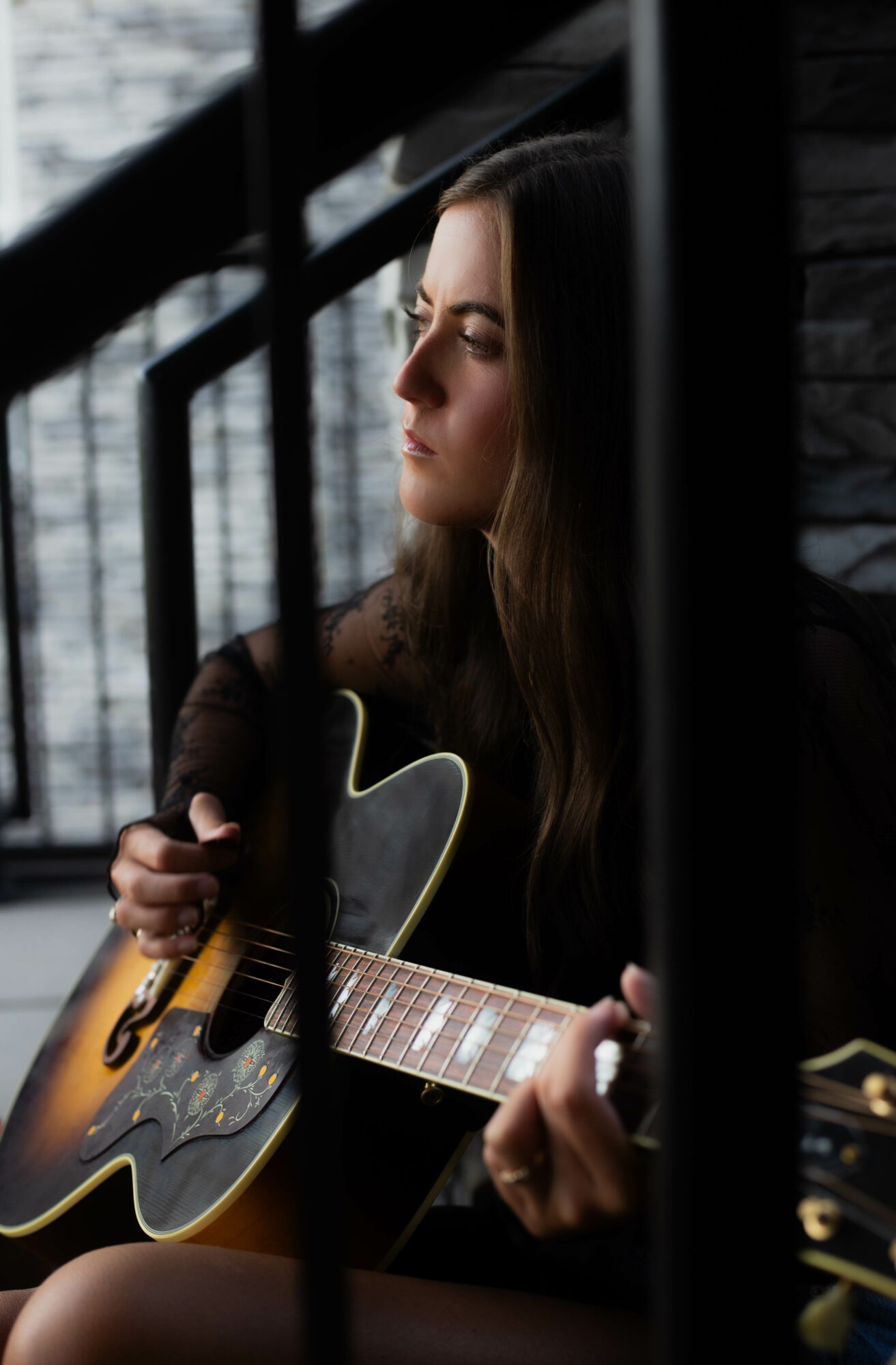 Woman with long hair playing an acoustic guitar, sitting near a window with black bars, looking contemplative, indoors.