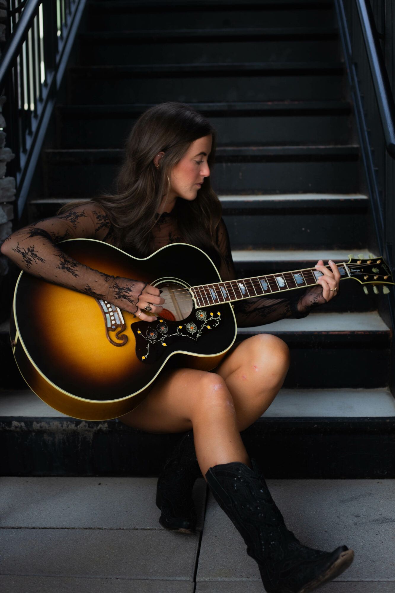 Woman sitting on stairs playing an acoustic guitar, wearing a black dress and boots, with dark hair.