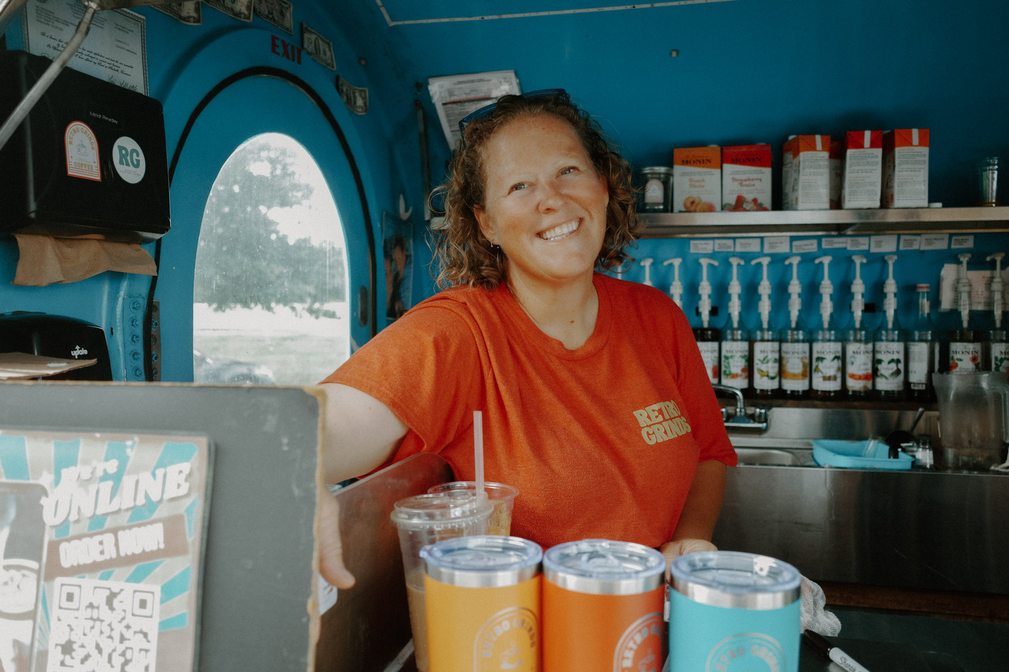 Smiling woman with curly hair in orange shirt behind a counter with drinks and supplies inside a colorful cafe or shop.