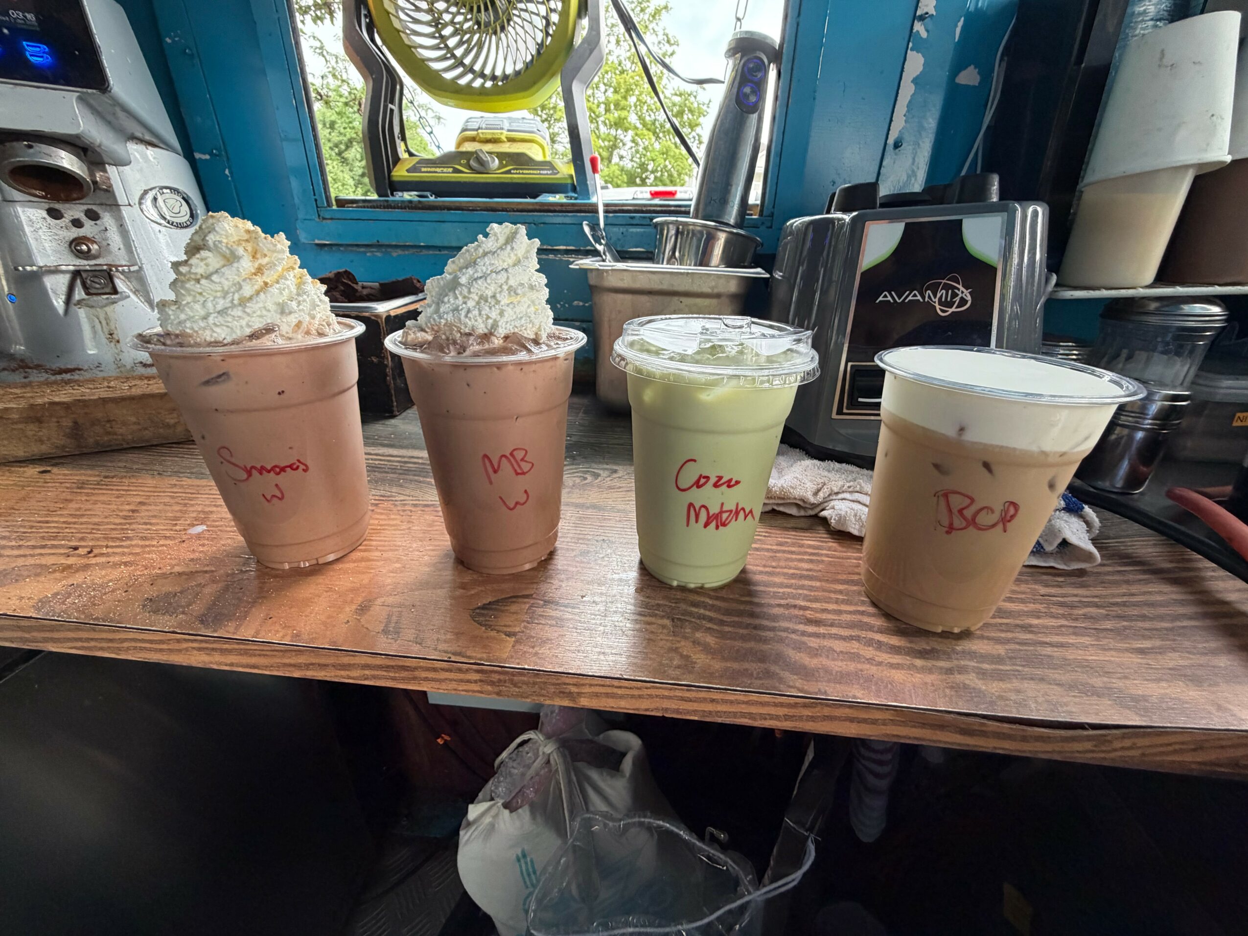 Four beverages on a wooden counter, topped with whipped cream or foam, in a cozy cafe setting.