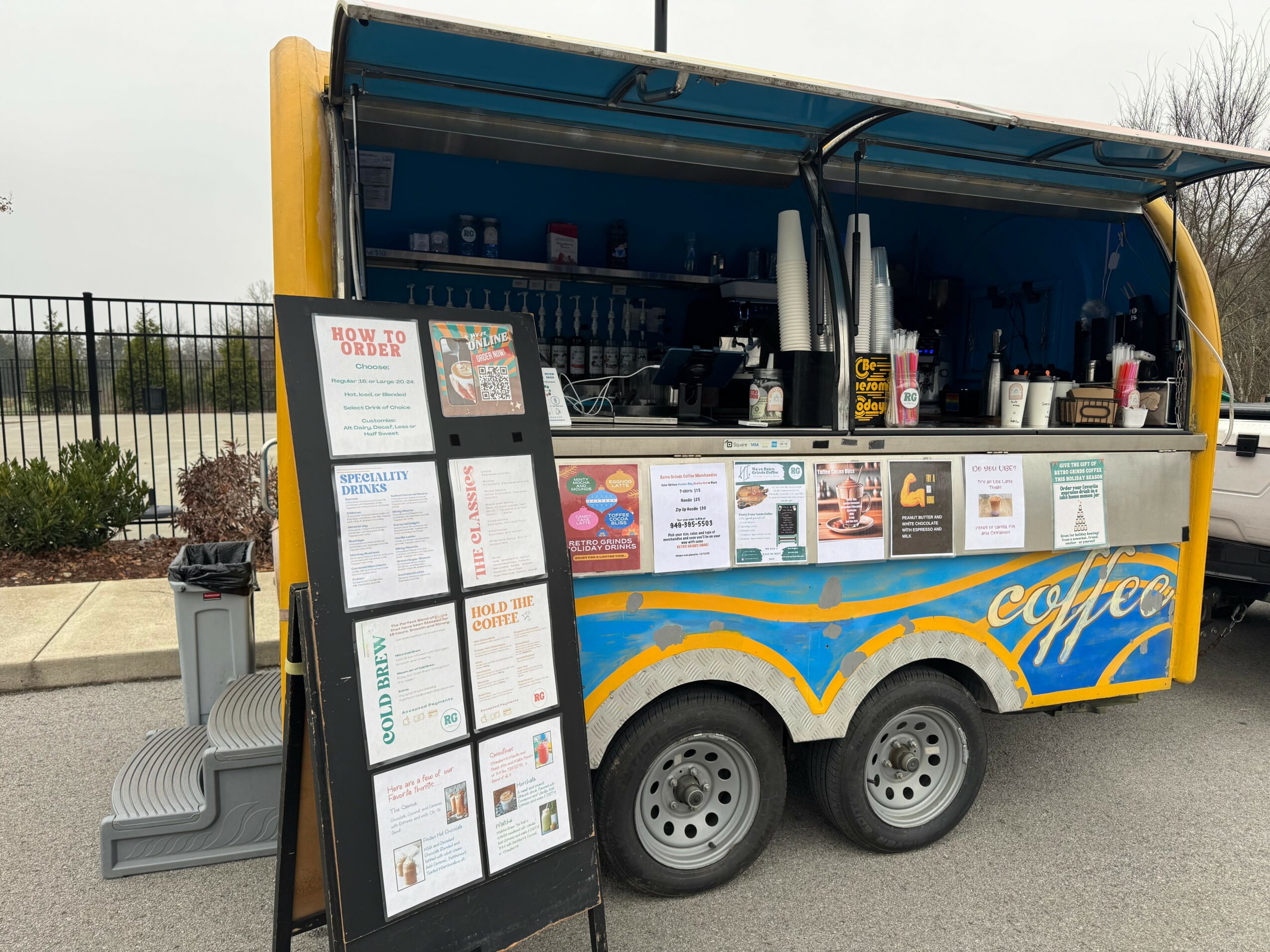 Mobile coffee cart with menu boards, serving area, and wheels, parked outdoors with a fence and trees in background.