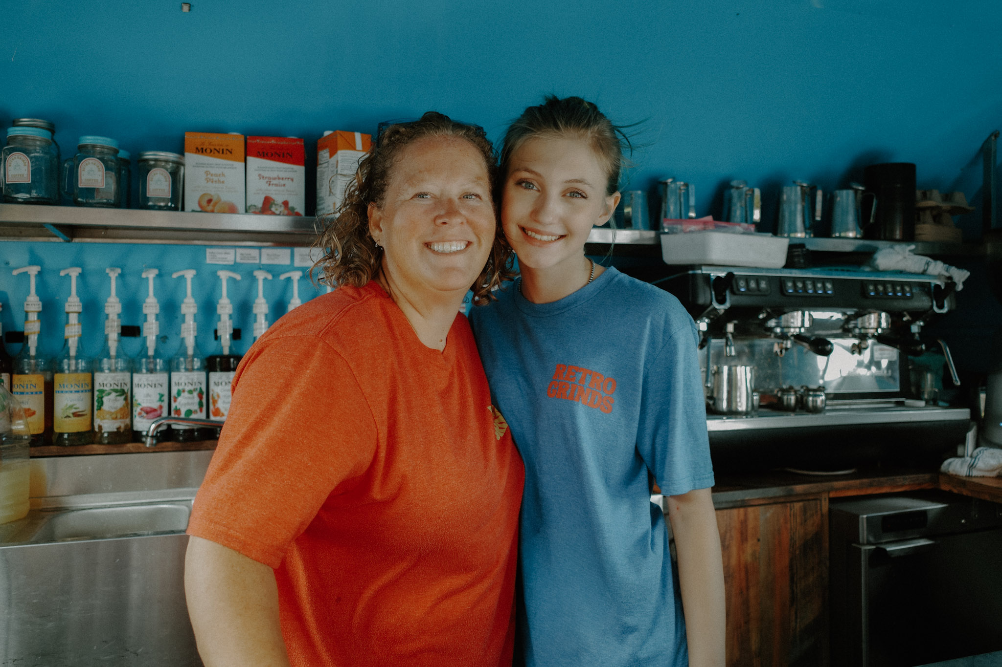 Two women smiling in a coffee shop, one wearing a red shirt and the other a blue shirt, with coffee equipment behind them.