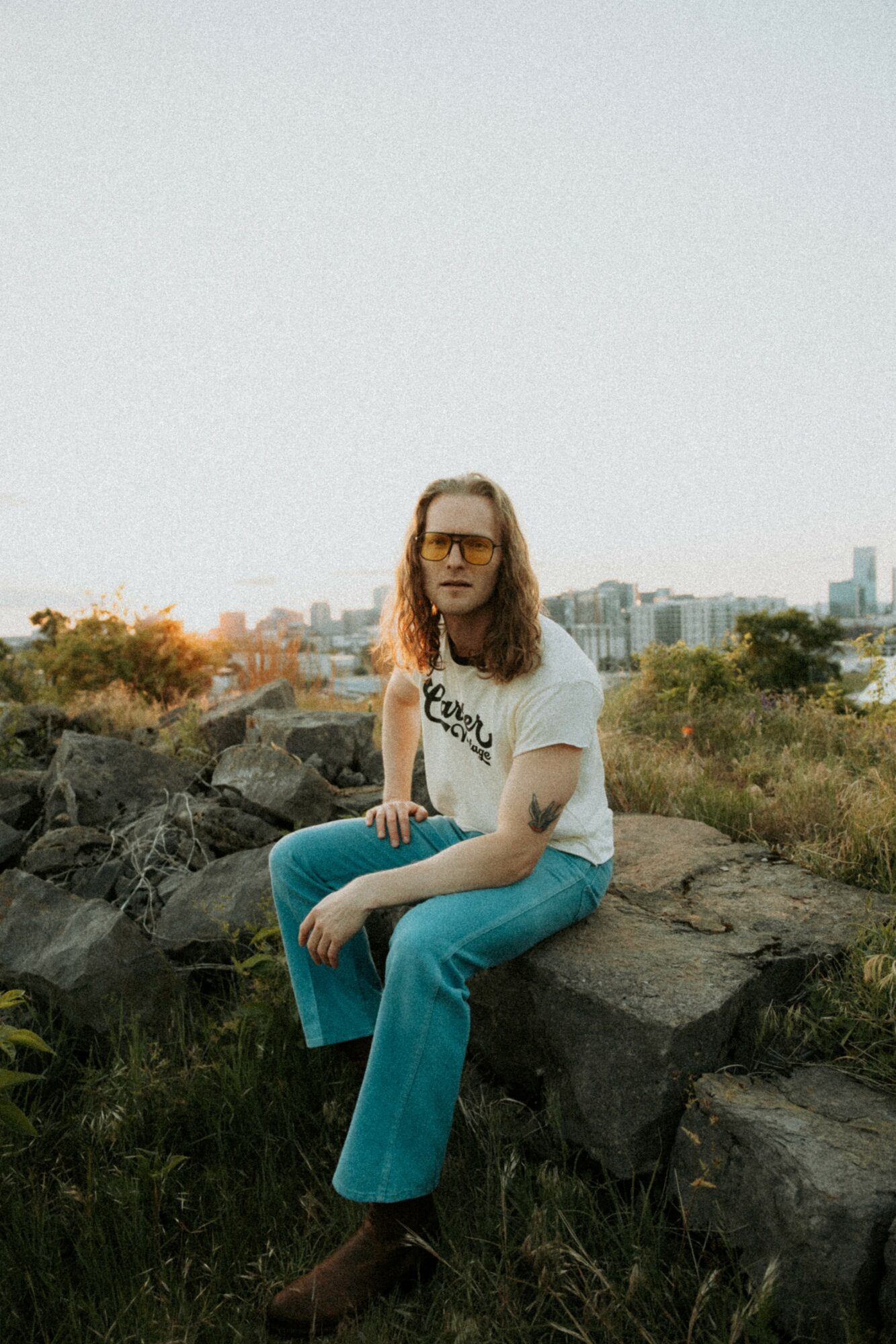 Person with long hair and glasses sitting on rocks outdoors, city skyline in background, daytime.