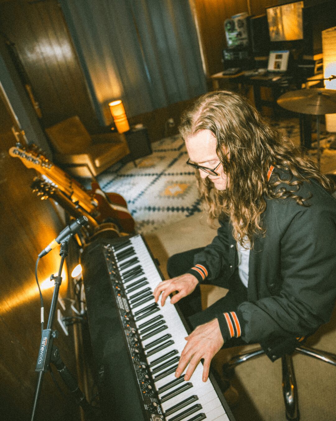Person with long hair and glasses playing a keyboard in a cozy room with music equipment and warm lighting.