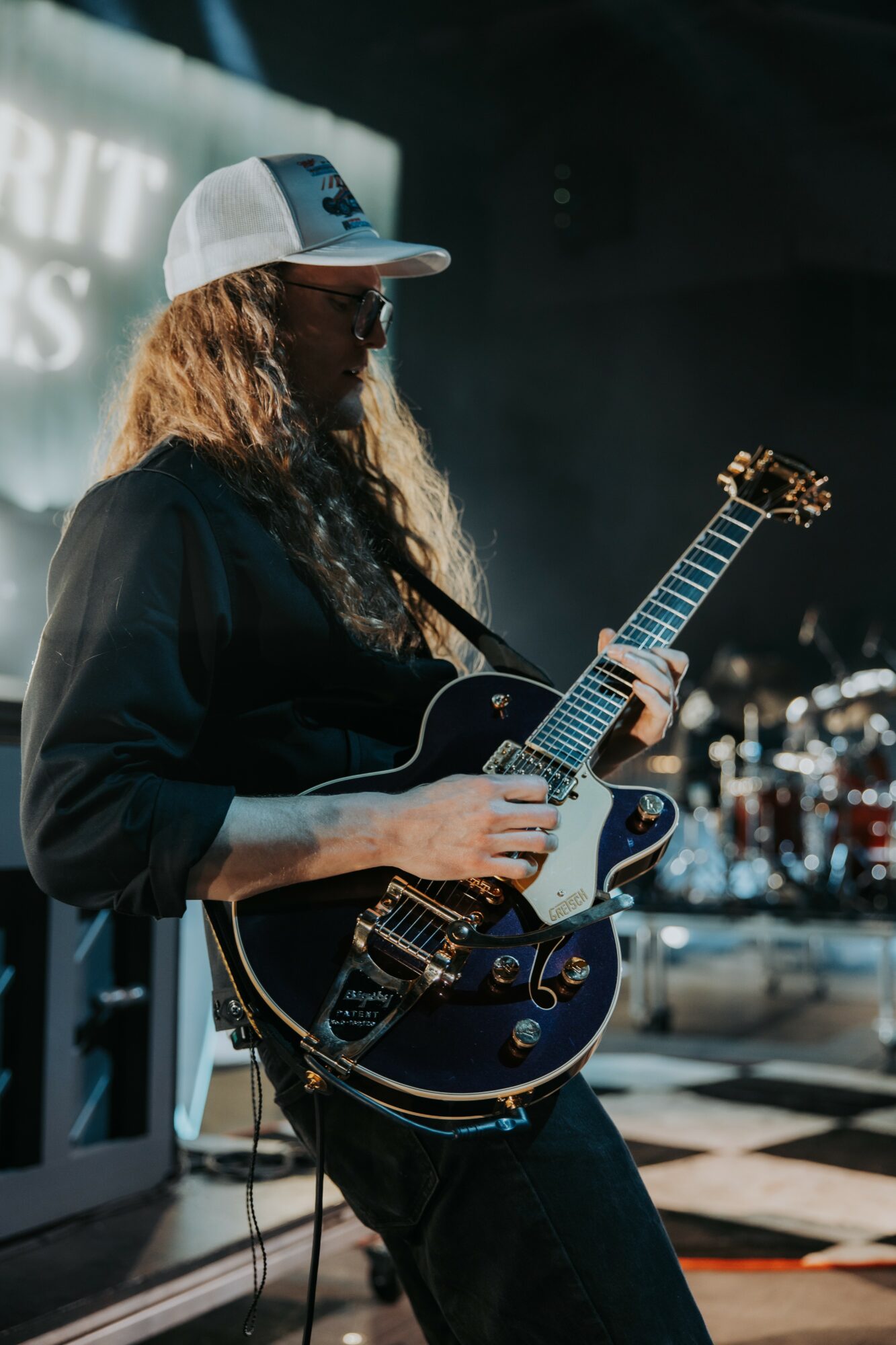 Person playing an electric guitar on stage, wearing a cap and dark clothing, with a drum set in the background.
