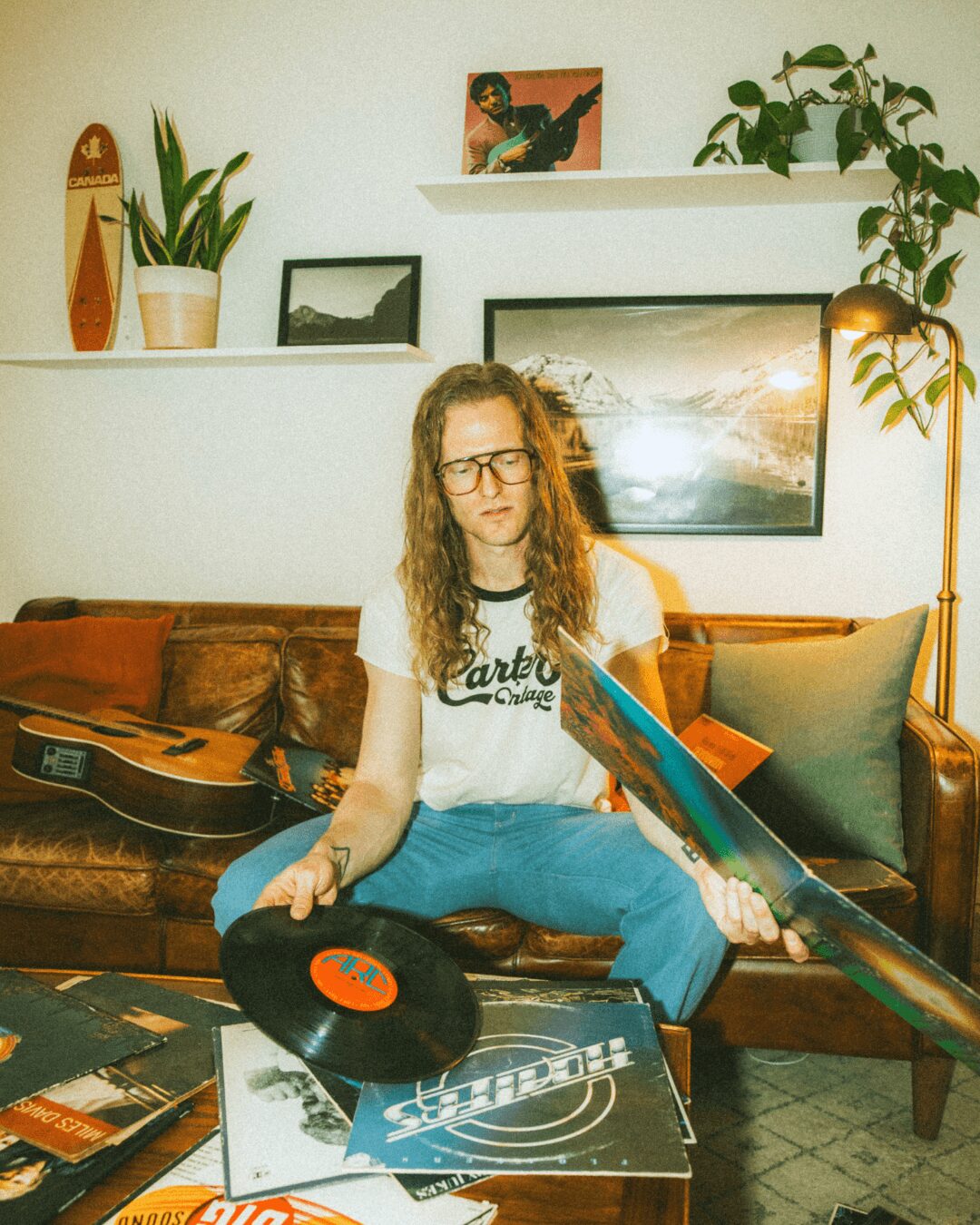 Person with long hair and glasses sitting on a sofa, holding a vinyl record and a guitar, surrounded by plants and artwork.