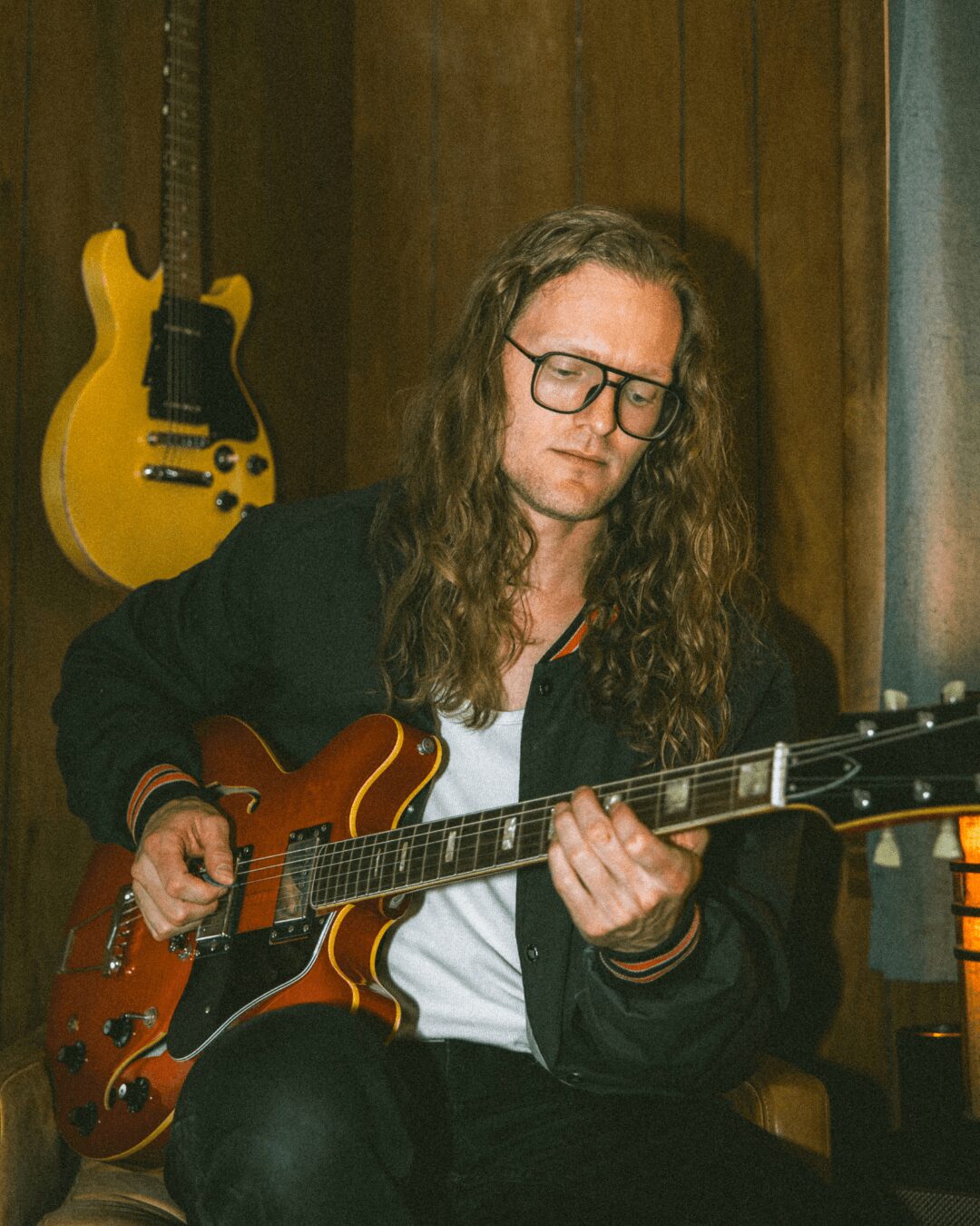 Person with long hair and glasses playing an electric guitar indoors, with a yellow guitar hanging on the wall behind.
