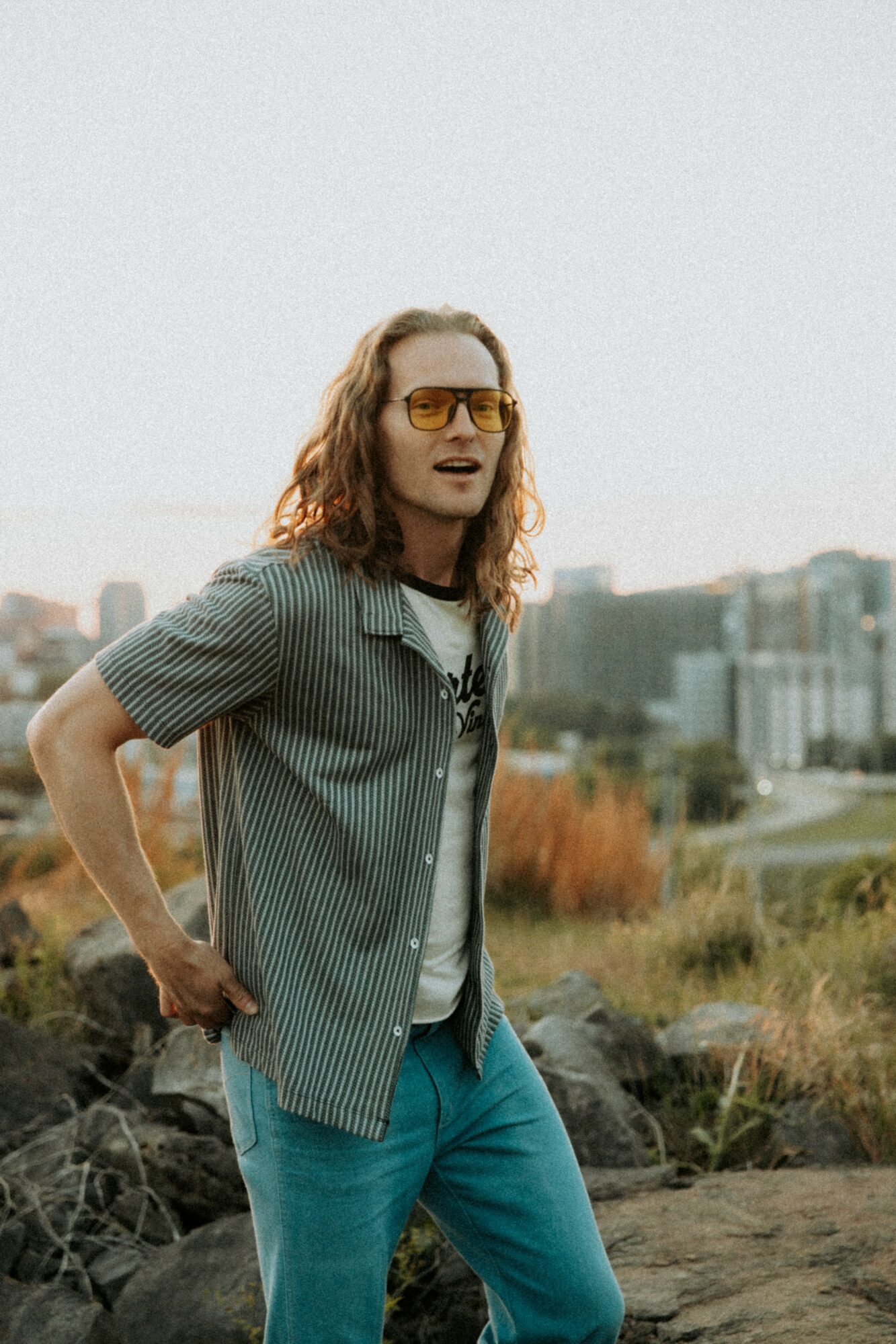 Person with long hair wearing sunglasses, striped shirt, and jeans outdoors with cityscape background.